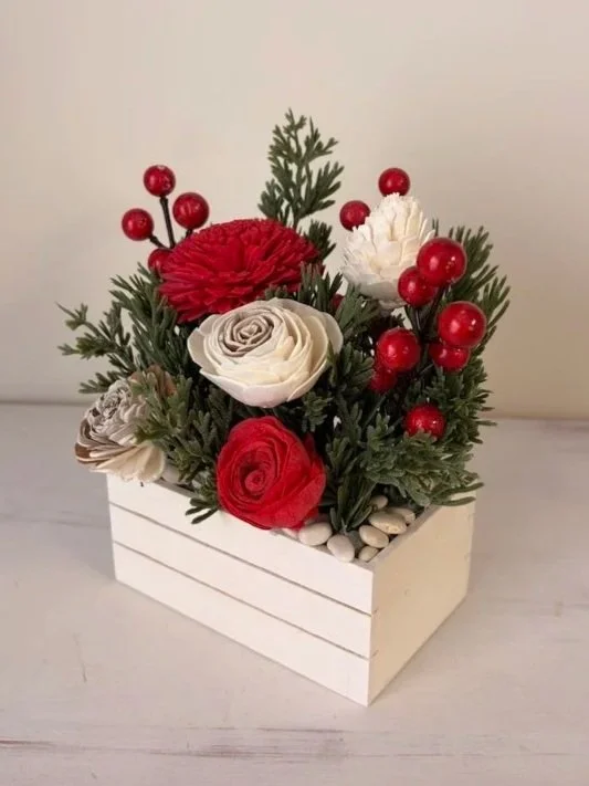 Decorative flower arrangement with red and white flowers, green foliage, red berries, and white stones in a white wooden box.