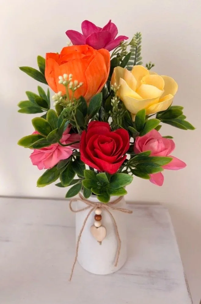 Colorful artificial flower bouquet with roses, tulips, and greenery in a white ceramic vase with a twine bow and small heart decoration.
