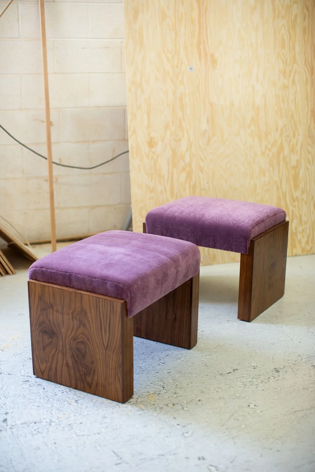 Two wooden handcrafted stools with purple upholstered cushions are placed on a concrete floor, against a unfinished wooden wall.