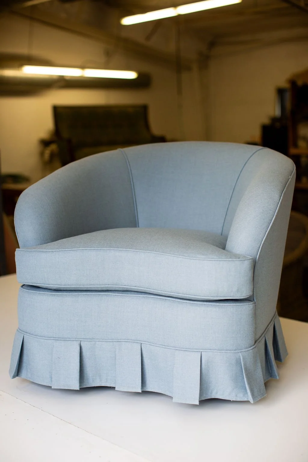 Light blue upholstered armchair with box pleated skirt at the base in a room with dim lighting and dark furniture in the background.