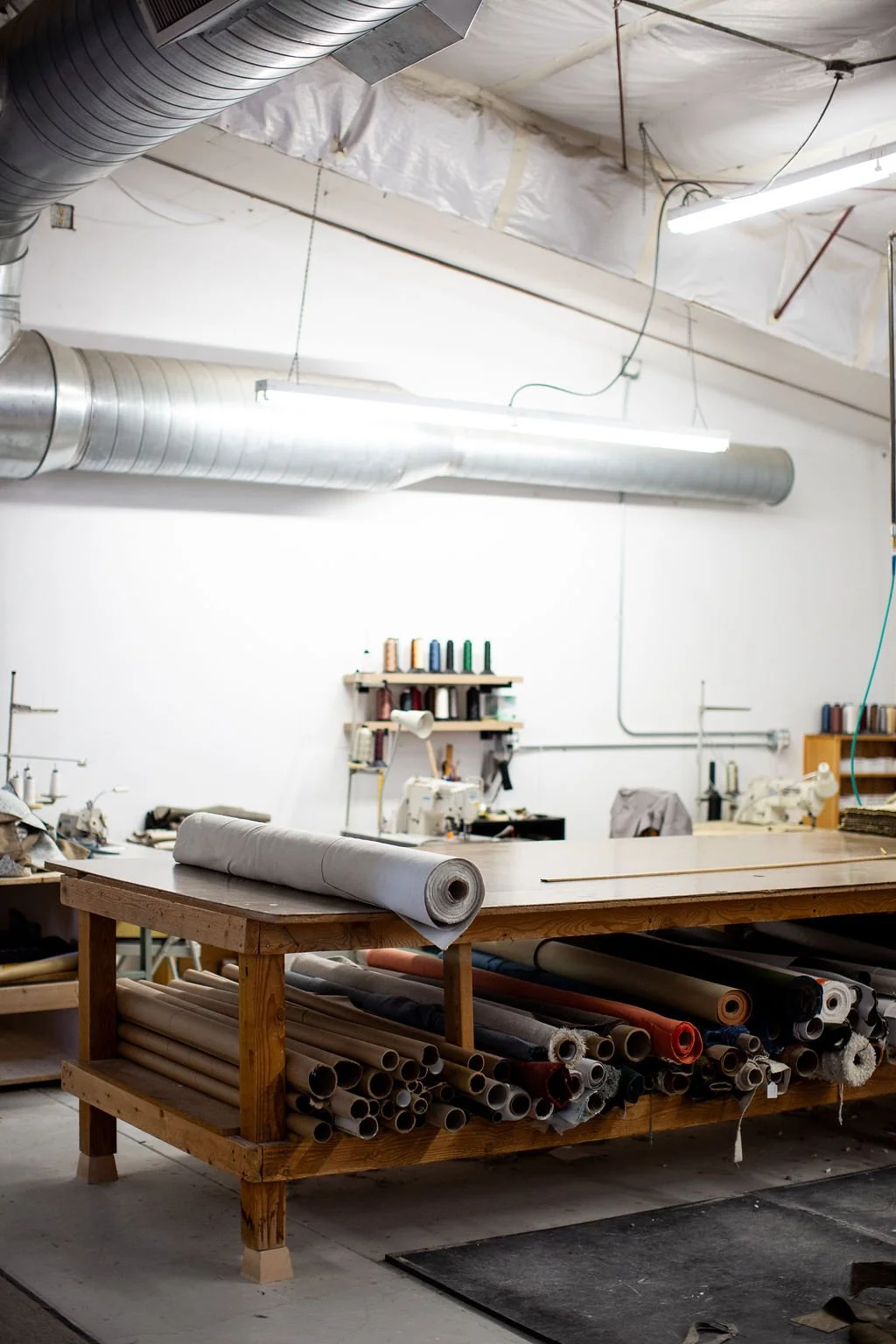 A furniture upholstery workshop with a large wooden table holding a roll of fabric, surrounded by shelves filled with various rolls of fabric, and sewing supplies. The ceiling has exposed ductwork and is unfinished with insulation and hanging lights.