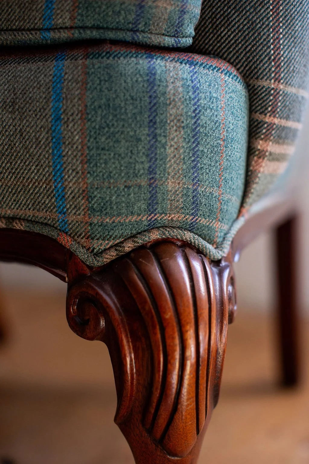 Close-up of a wooden furniture leg with curved carvings, above it a green plaid pattern fabric upholstered to the chair, divided by double welt in green plaid fabric.