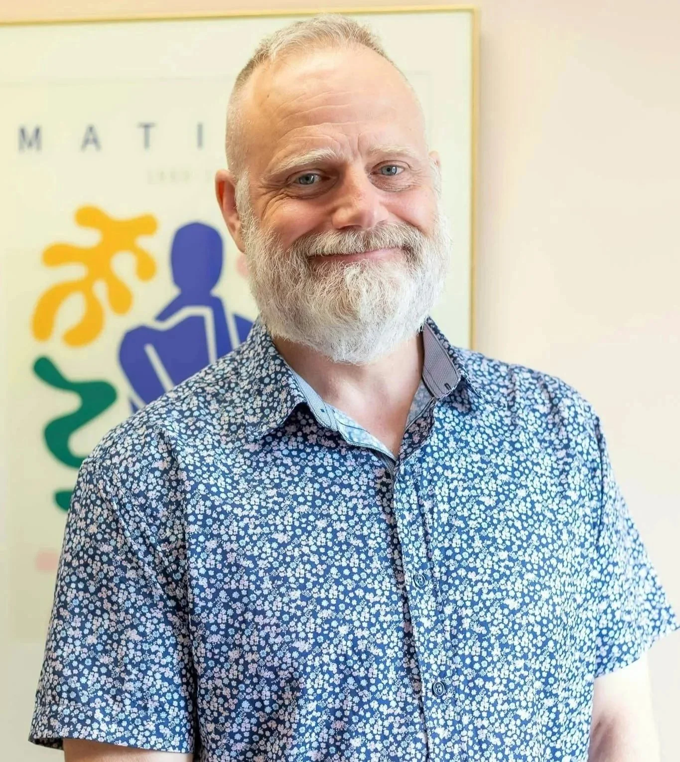 Chris Jackson, smiling with a beard and mustache, wearing a patterned short-sleeve shirt, standing in front of a colorful poster with abstract human-like figures.