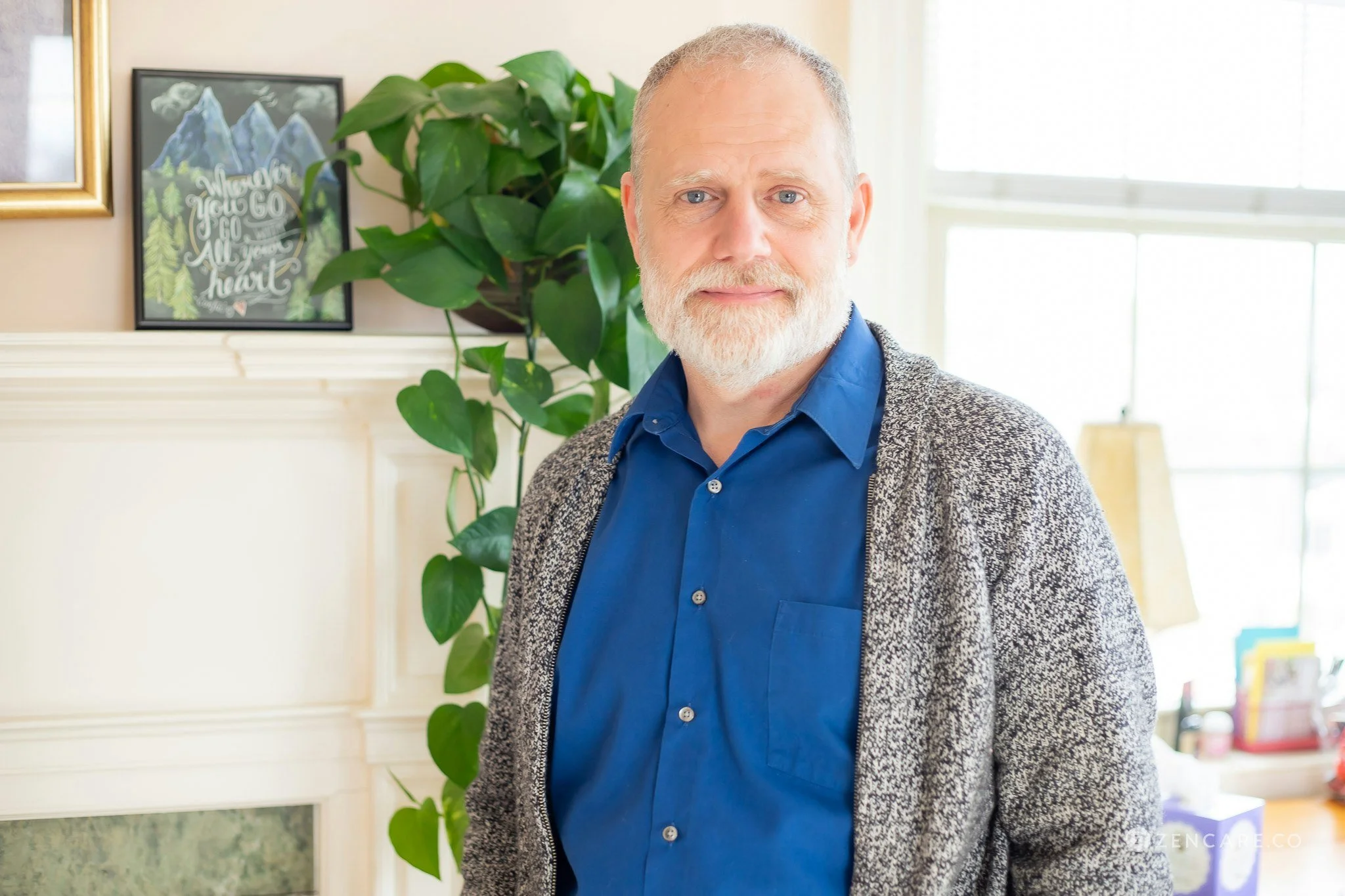 Chris Jackson with a beard in a cardigan and blue shirt smiling in front of a plant.