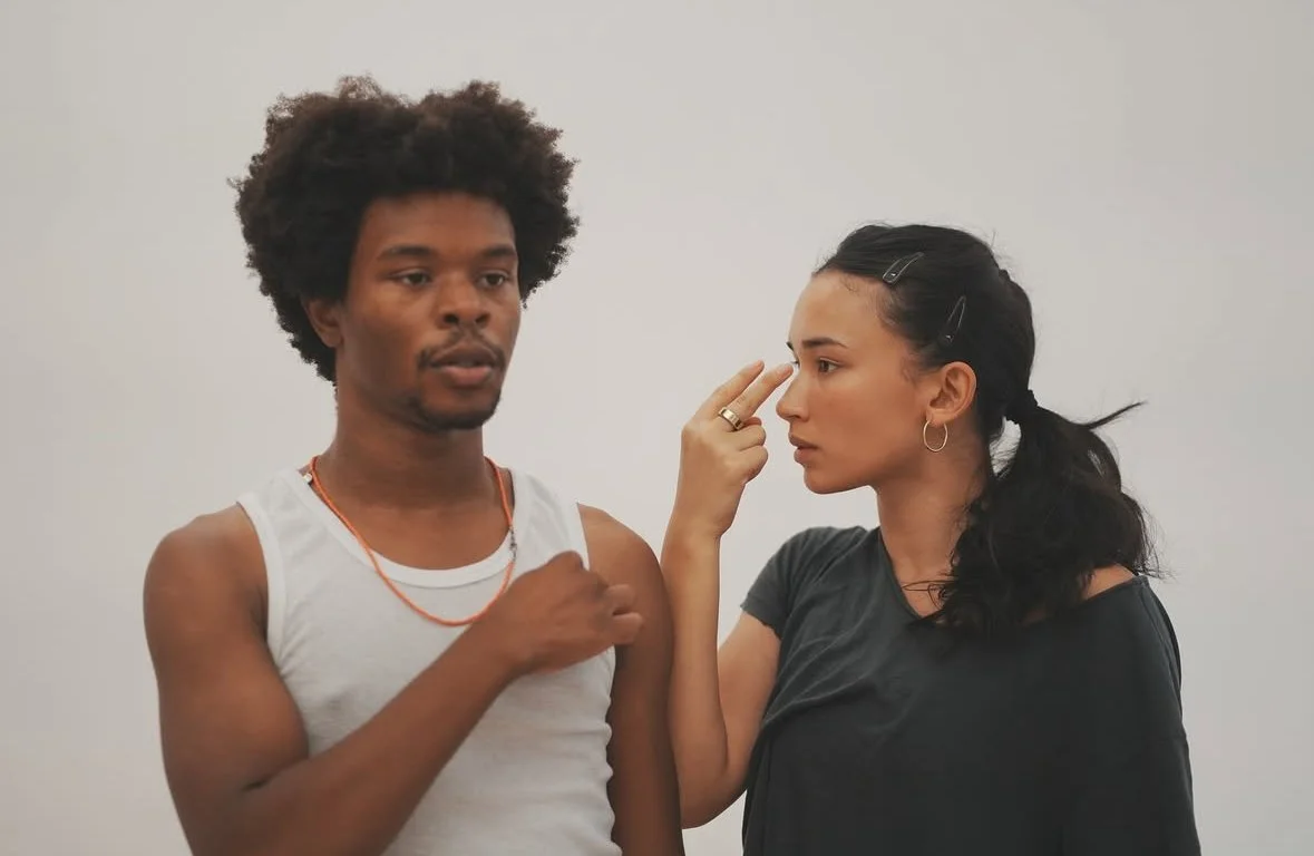 A young man with curly hair in a white tank top looks at a woman with dark hair, who is touching her nose with her finger and looking at him.