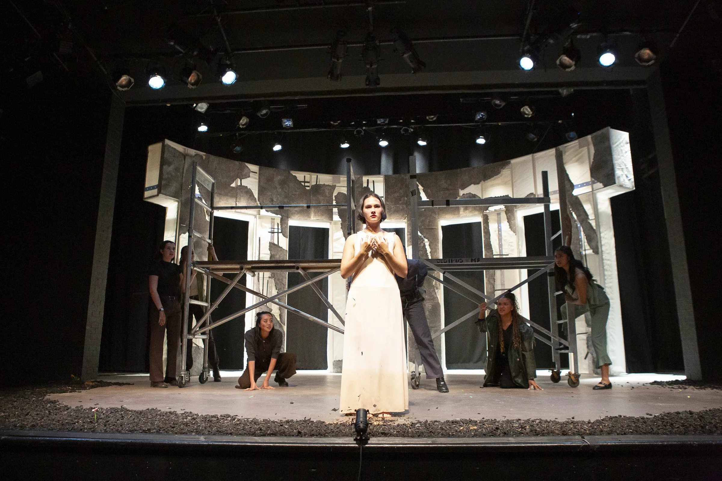A theatrical stage with five women in costume, one central woman in a long white dress standing with hands on her chest, surrounded by four women in casual or darker clothing, with a backdrop resembling a destroyed or old building, and stage lighting