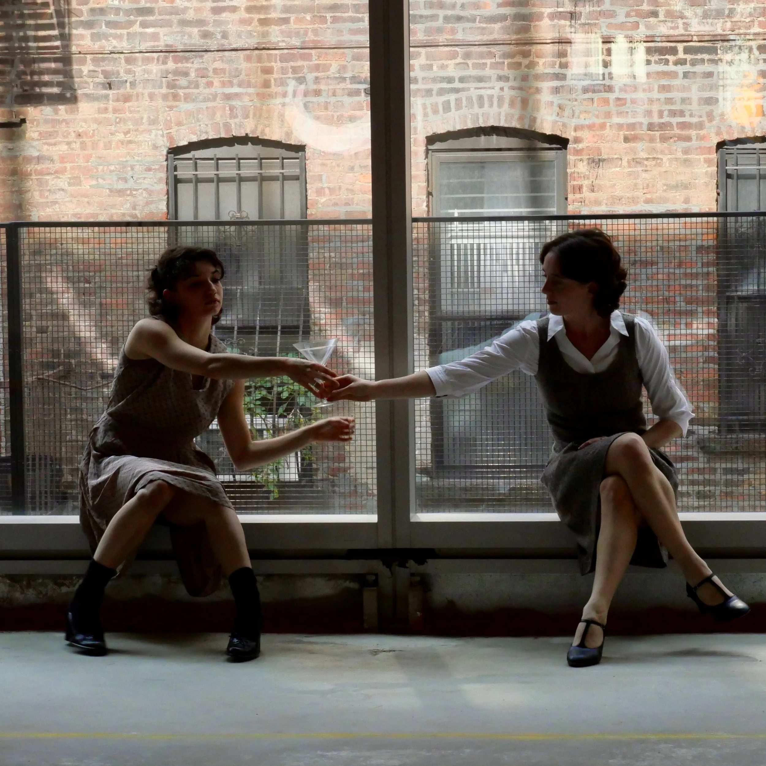 Two women sitting by a large window, passing a martini glass between them. The woman on the left wears a sleeveless dress, and the woman on the right wears a white blouse with a dark vest and skirt. The scene has an urban brick wall background.