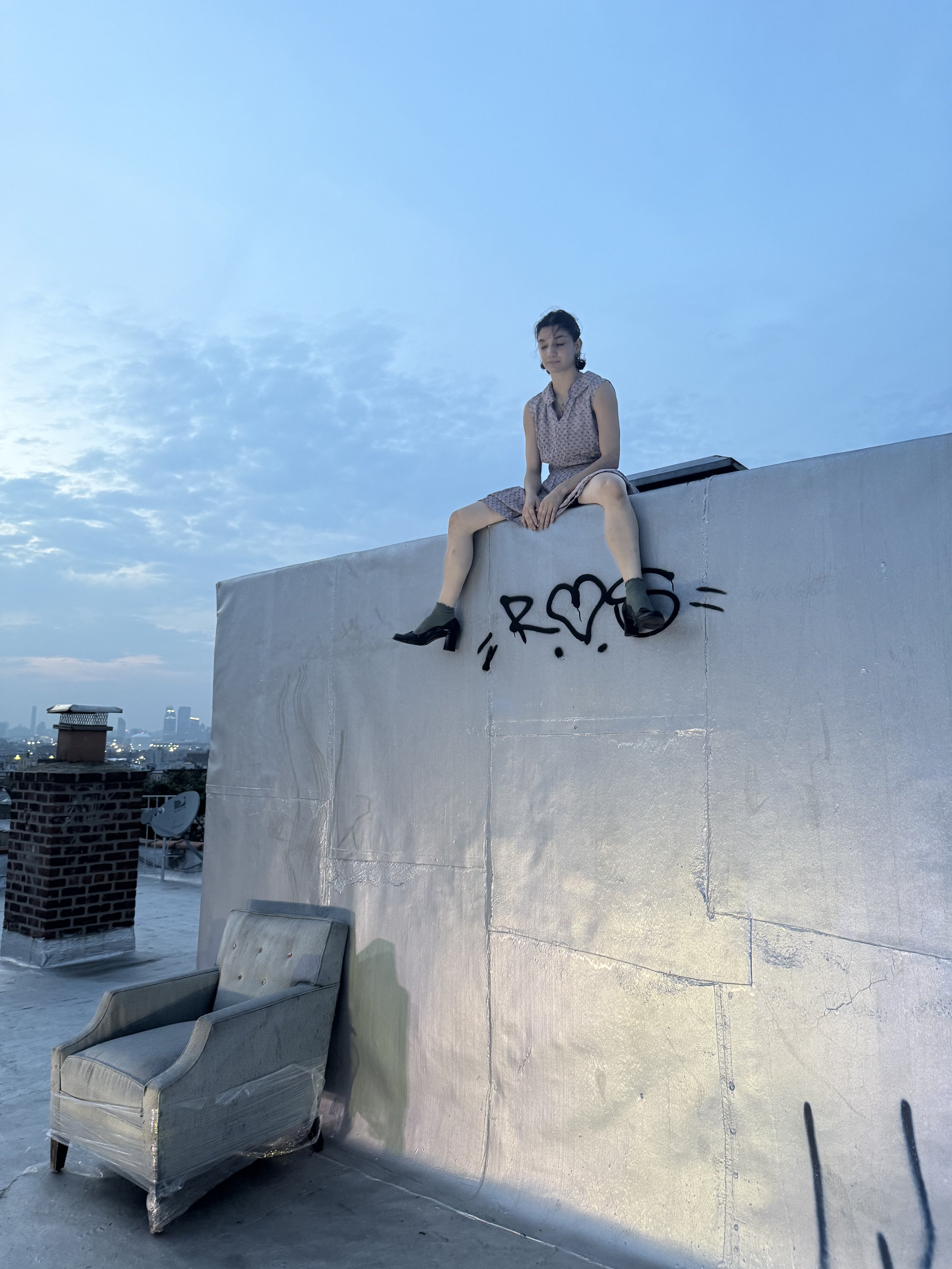 A young woman in a sleeveless dress and black shoes is sitting on the edge of a rooftop wall with her legs hanging down. The wall has graffiti with the word 'R' and a heart shape. A chair is placed on the rooftop below her, and the city skyline is vi