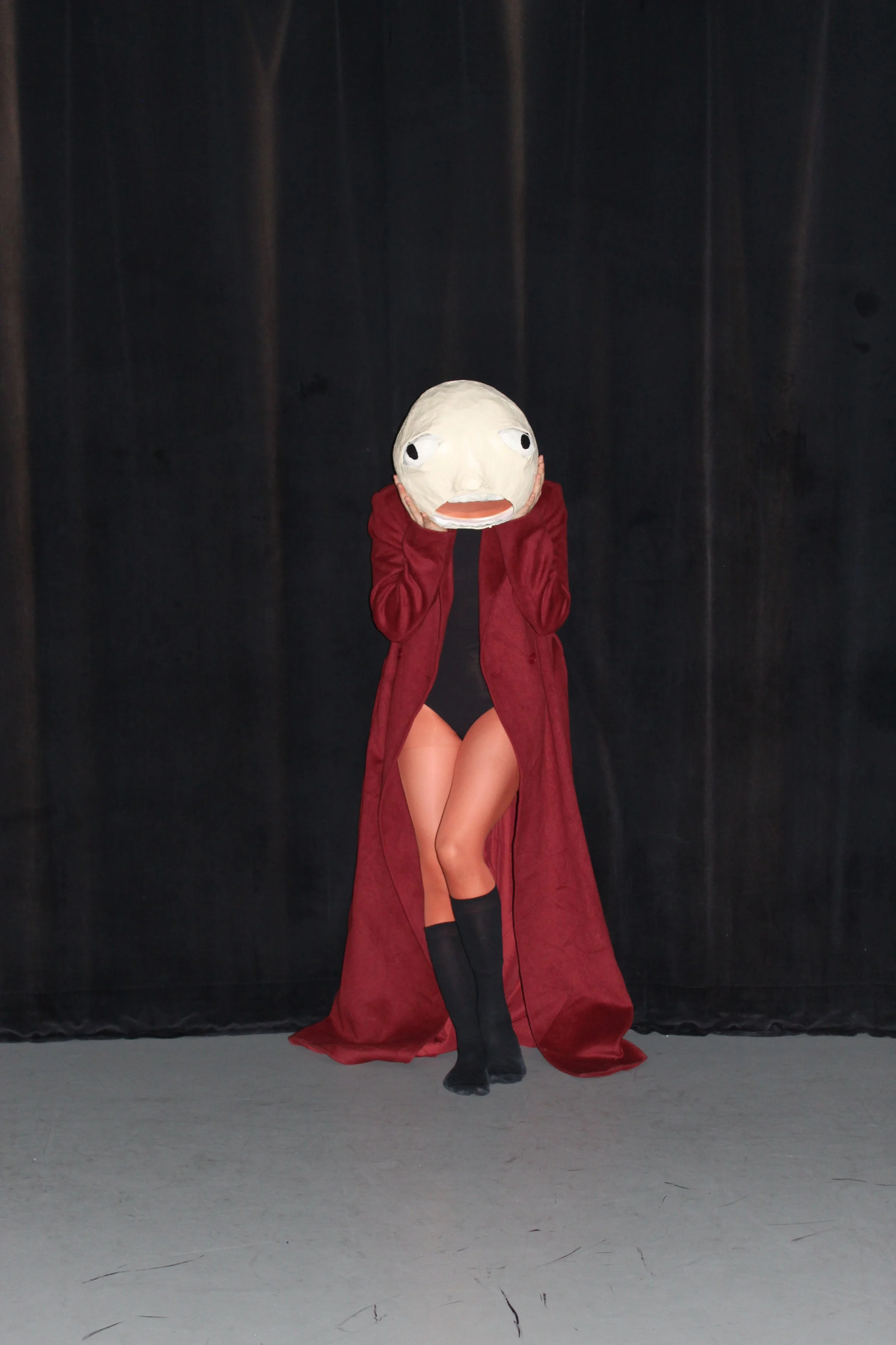 Person standing on stage wearing a mask with a large head and a long, red coat. They are holding the mask to their face with both hands, and they are wearing black boots.