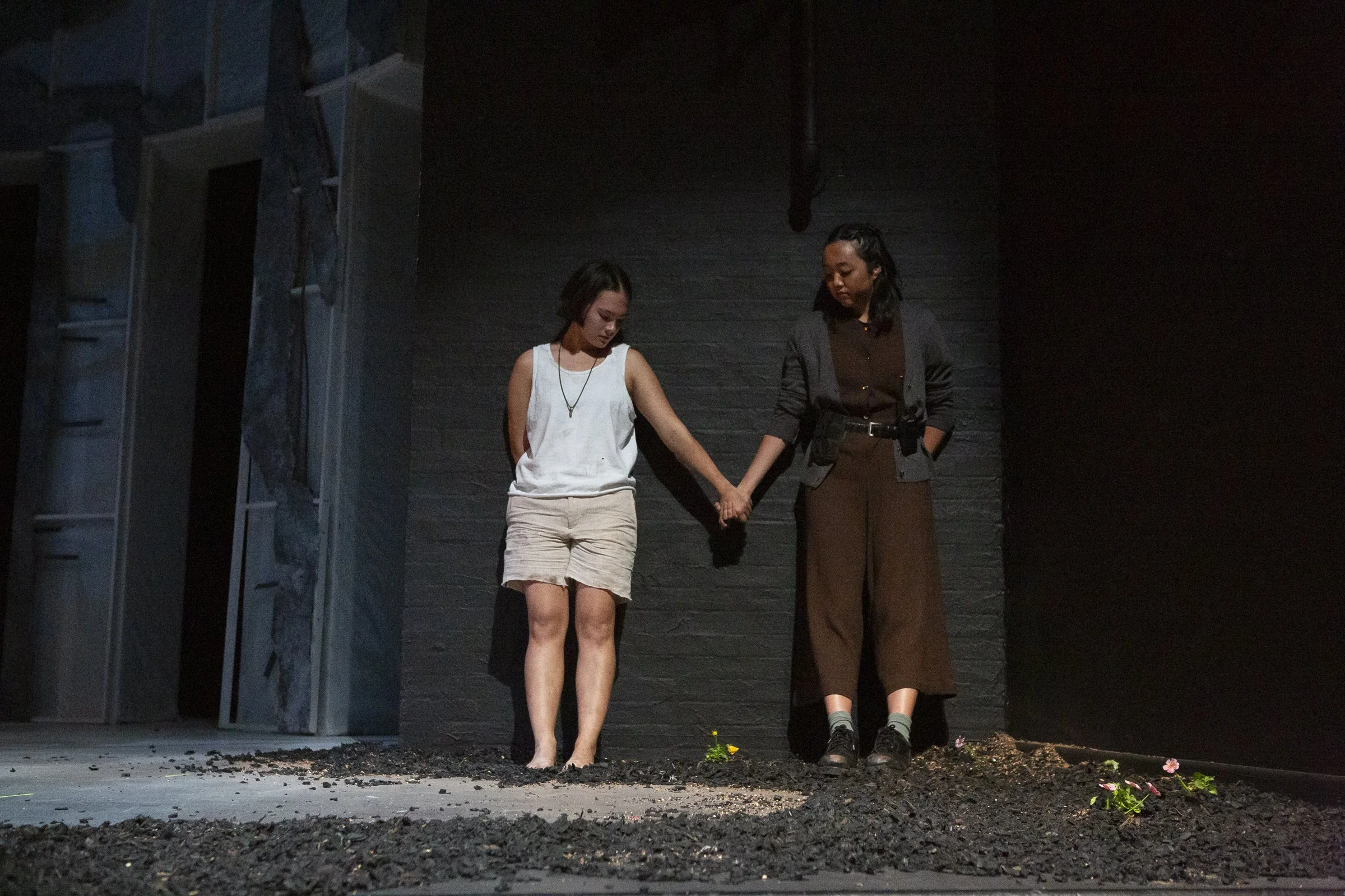 Two women hold hands and stand on black dirt in a dark room, with scattered flowers around them.
