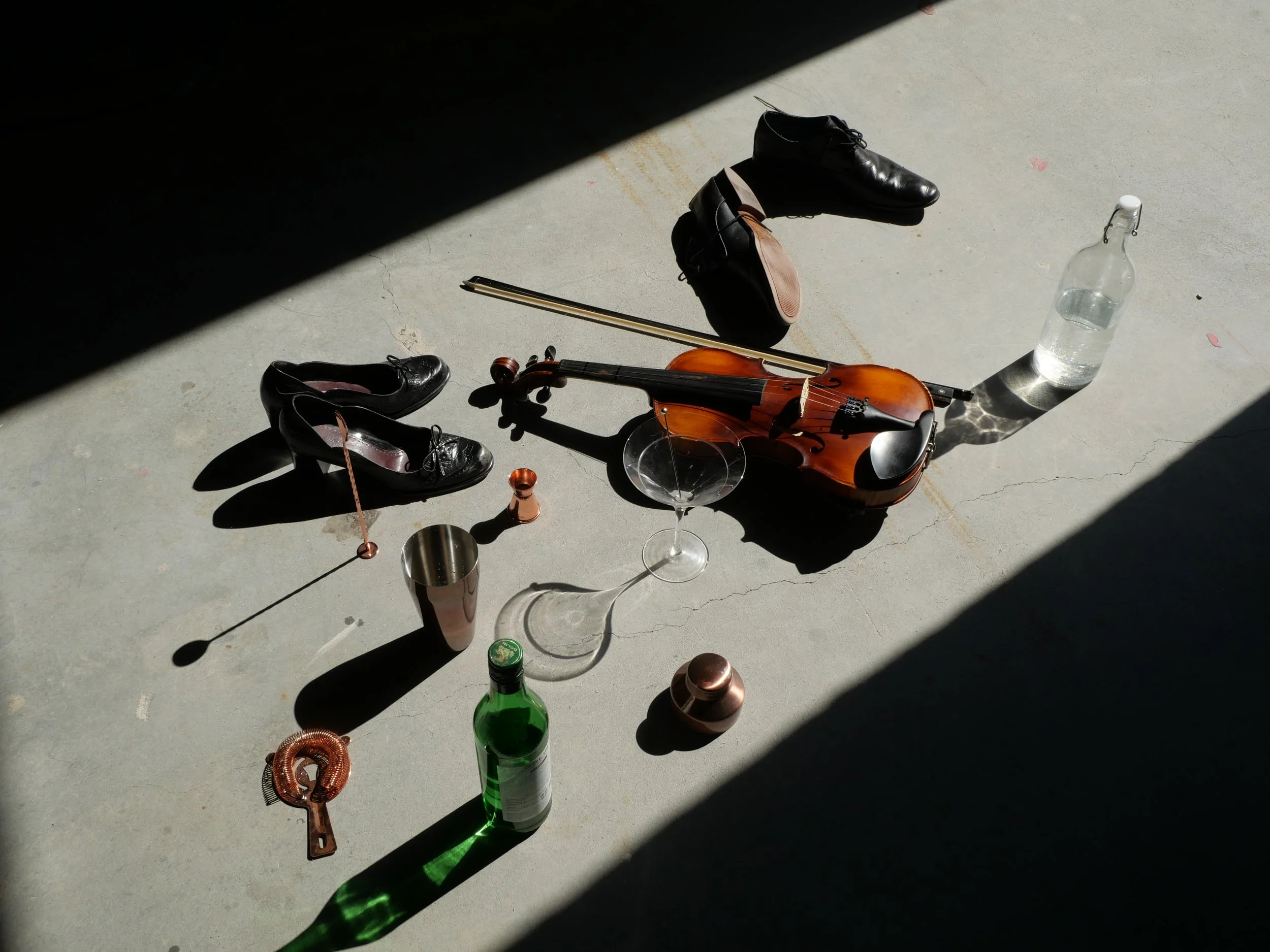 A violin with a bow, a pair of black shoes, a black handbag, a glass bottle, a wine glass, a green bottle, a copper key, and various small objects on a concrete floor with shadows and sunlight.