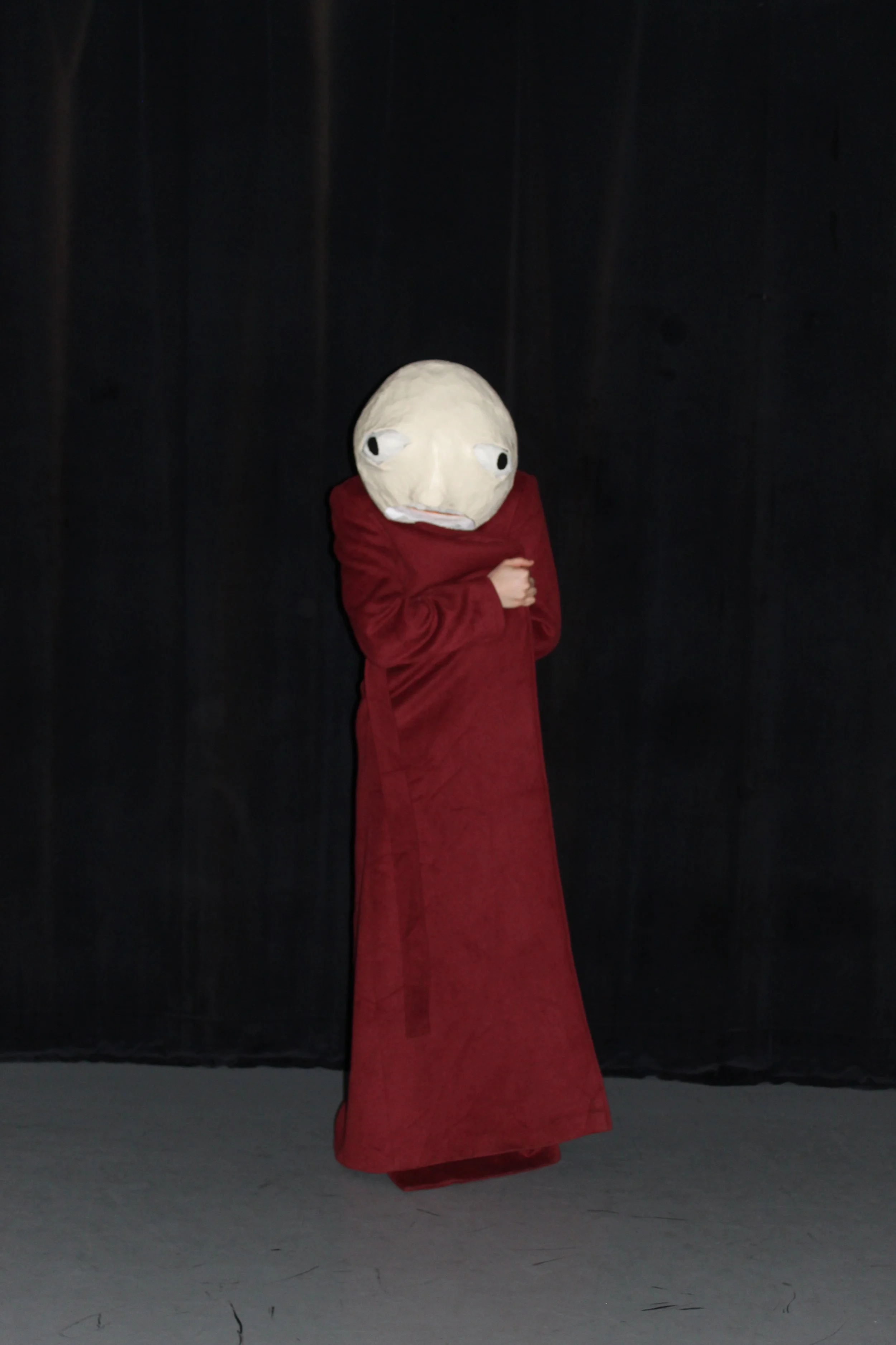 Person wearing a large, handmade puppet head with a neutral expression and eyes looking to the side, dressed in a long red dress, standing against a black curtain background.