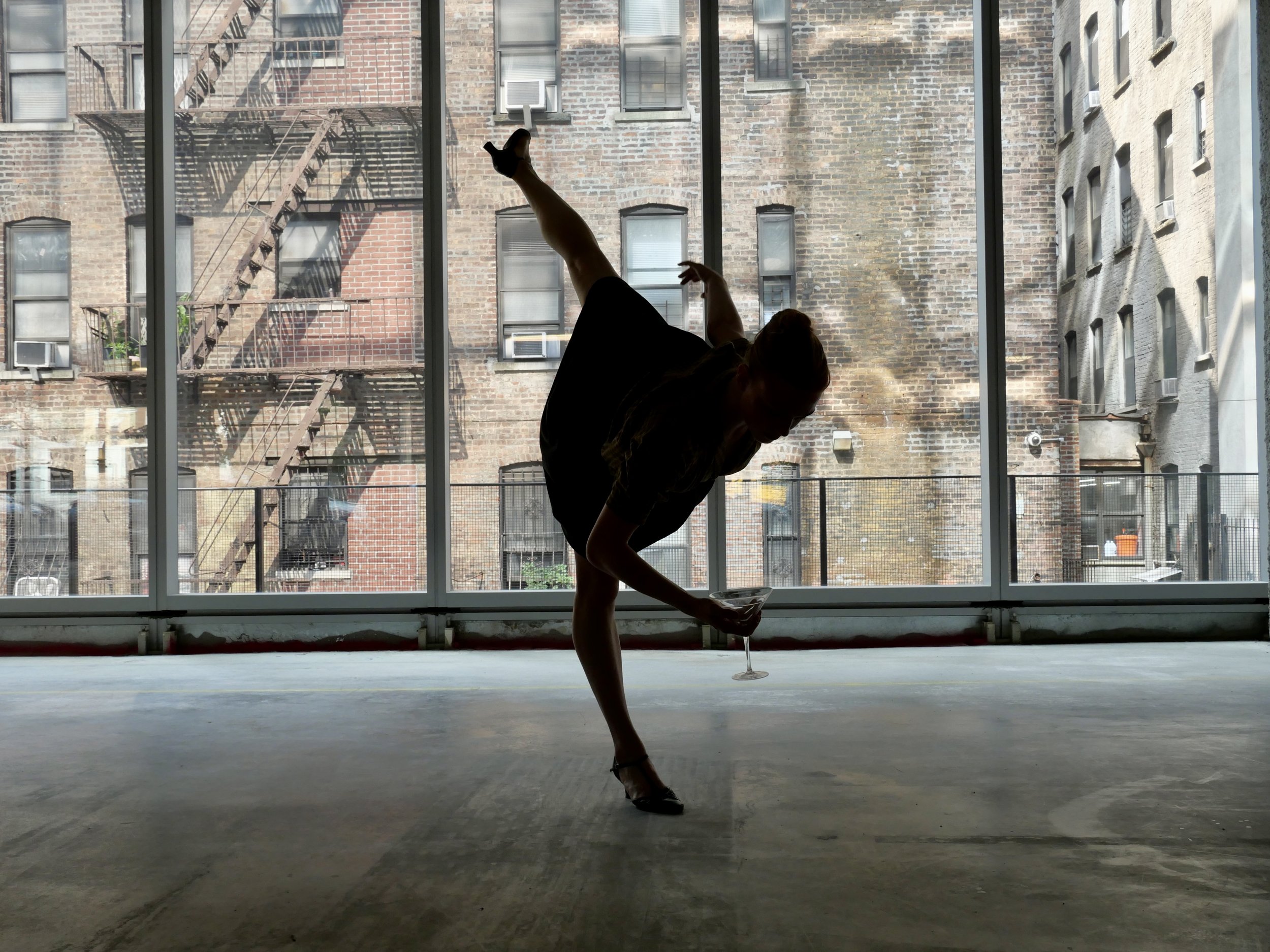 Silhouette of a woman holding a martini glass while performing a dance or gymnastic move in front of large glass windows with an urban cityscape background.