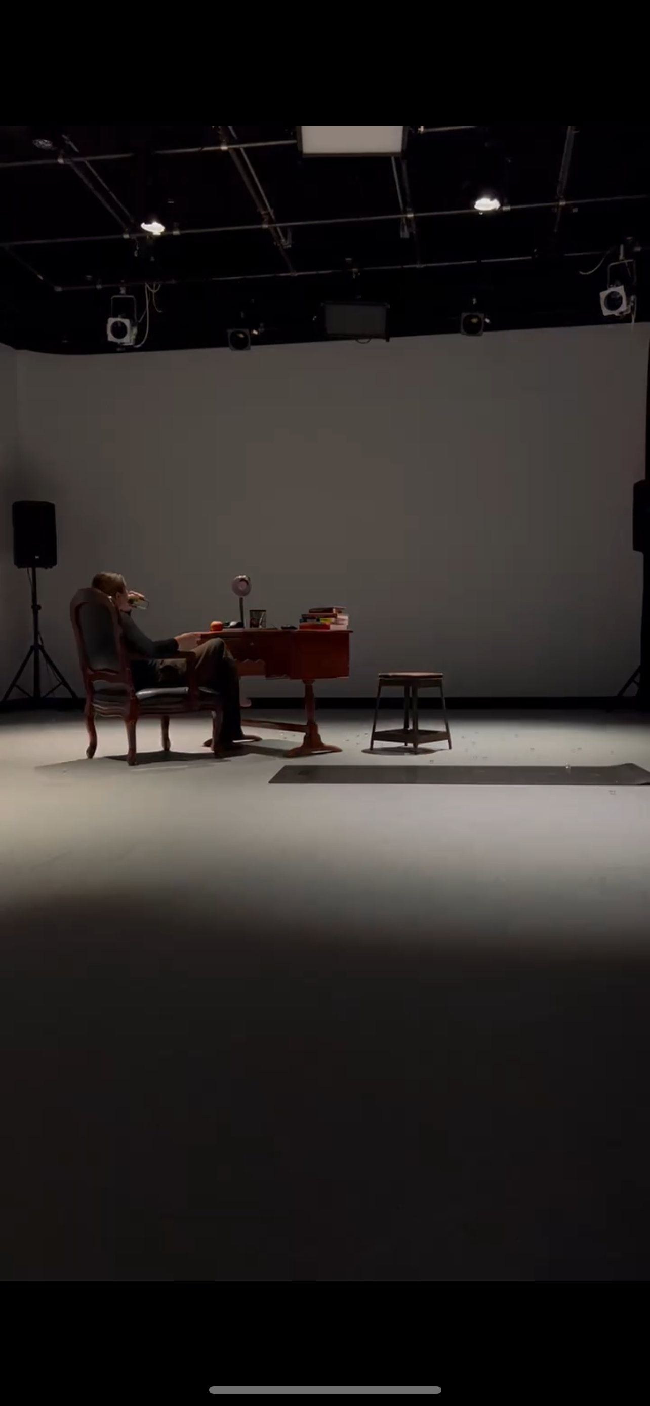 An empty stage with a man sitting at a wooden desk, drinking from a mug, surrounded by books, with a stool nearby, in a theater or studio setting.