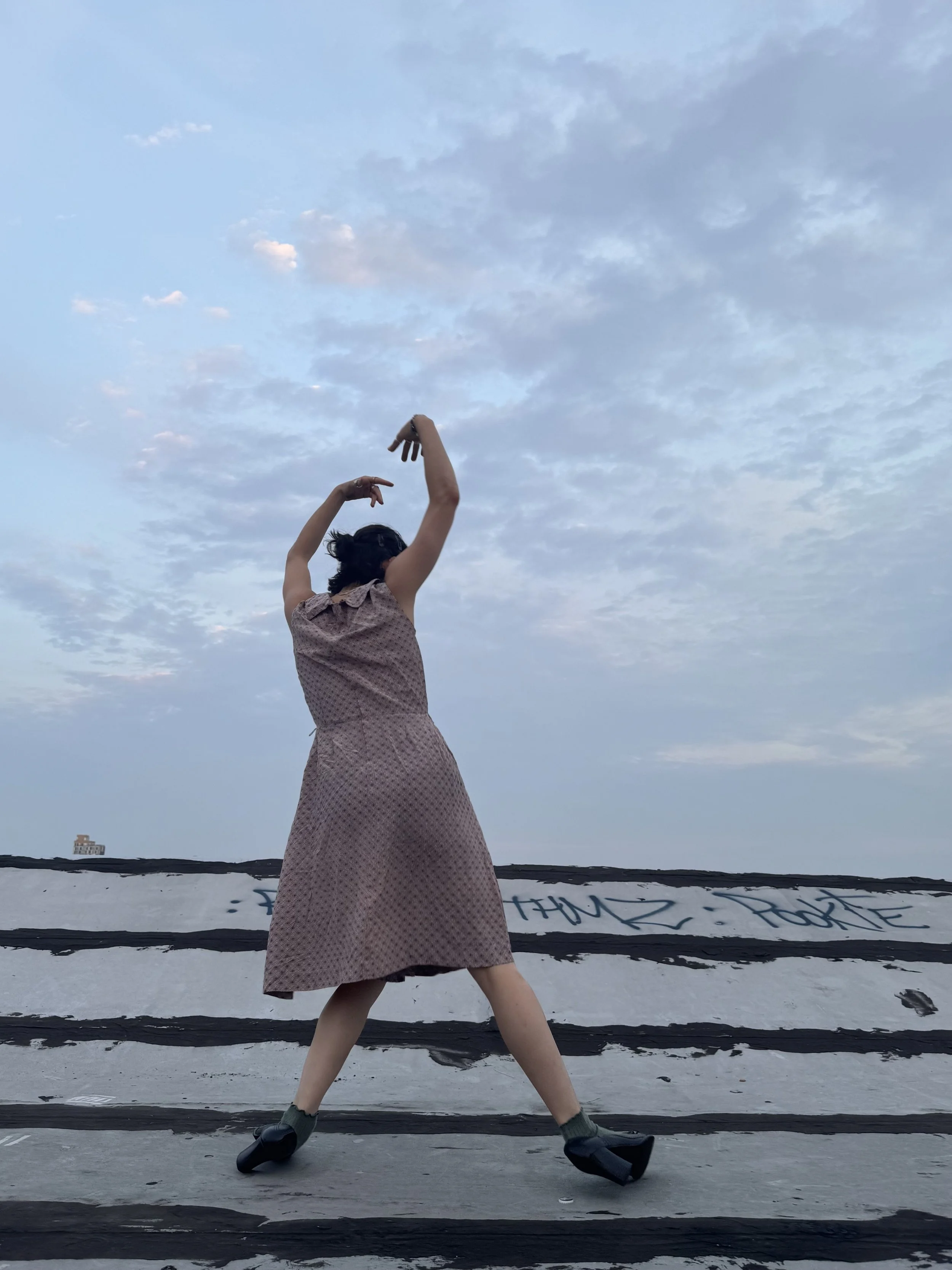 A woman standing outdoors on a rooftop or elevated area with graffiti, striking a dance pose under a cloudy sky.