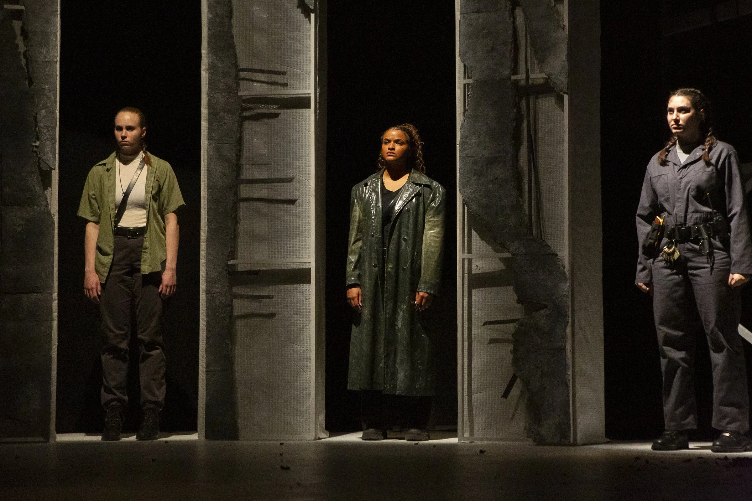 Three women standing in a theatrical stage set resembling prison or confinement cells, with dark background, illuminated from above, with somber expressions.