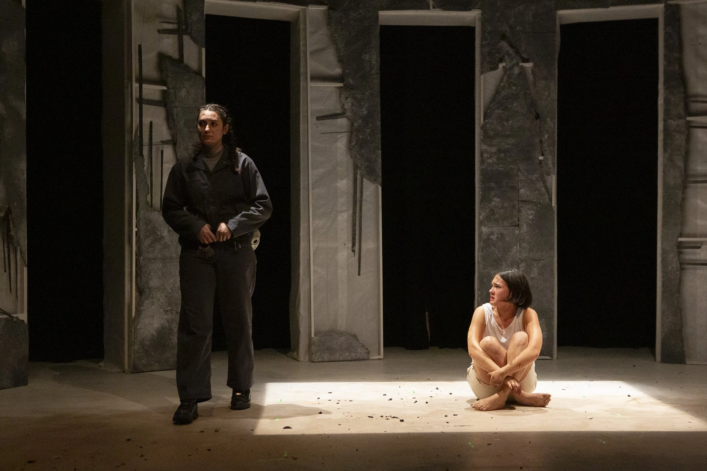 A theatre stage with two women acting, one standing and one sitting on the floor. The standing woman is dressed in black, and the sitting woman is in a white sleeveless shirt and beige pants, looking up at the other.