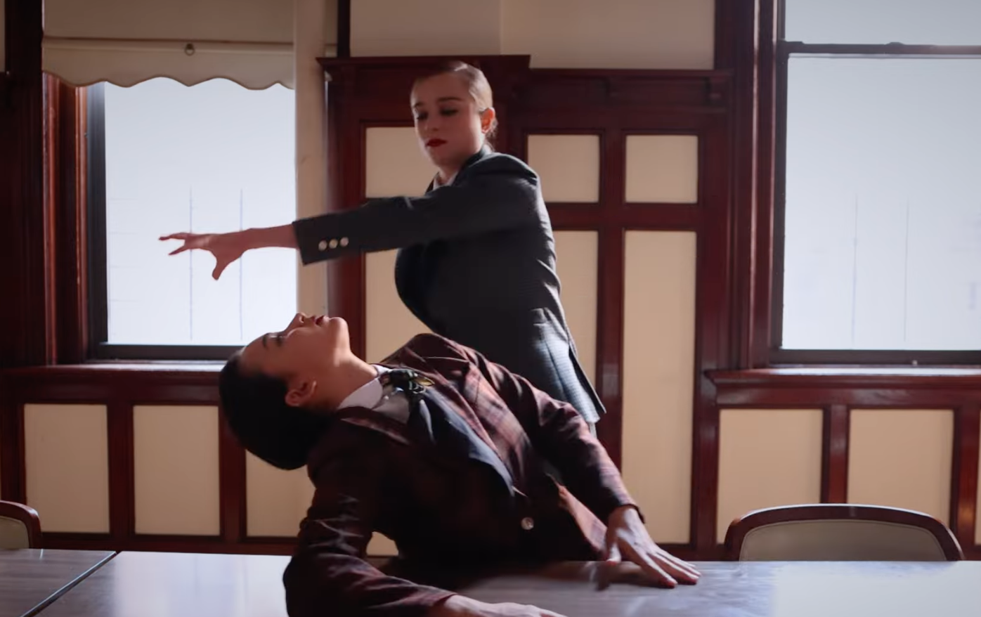 Two women in business attire performing a dance or stunt in a room with wooden paneling and large windows.