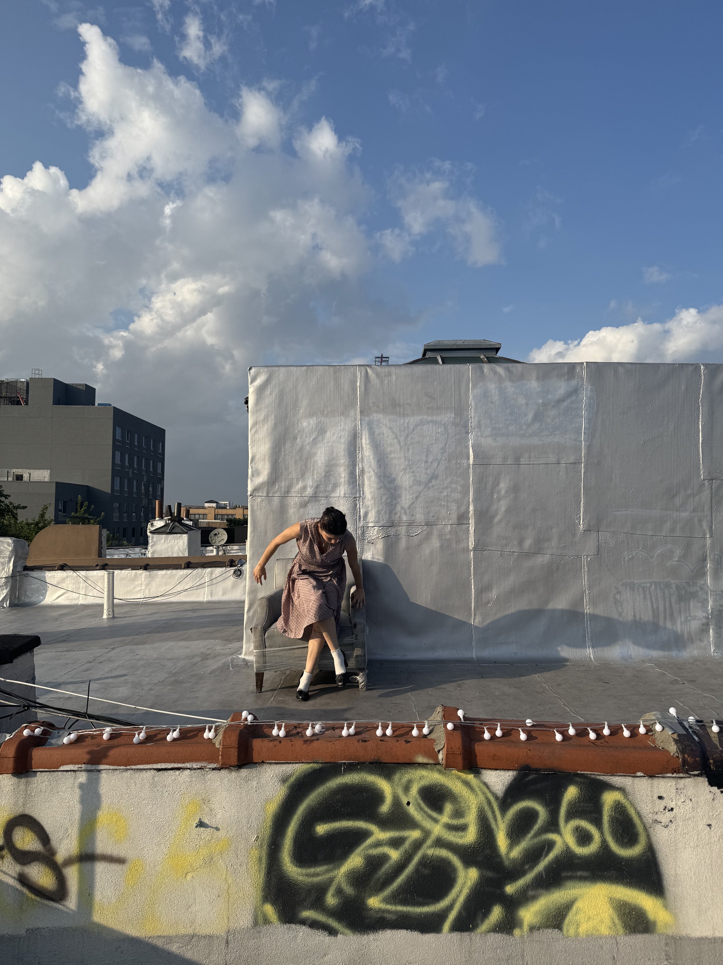 A woman in a patterned dress and white socks sits on an outdoor chair on a rooftop, adjusting her shoe. The rooftop has a gray surface, a white wall, and some graffiti at the bottom. The sky is partly cloudy with blue and white clouds.
