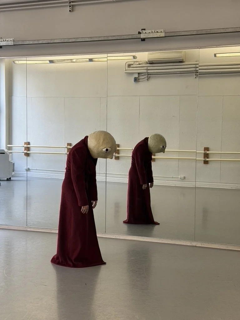 Person in a maroon dress wearing a large, round, beige headpiece with eyes, standing in front of a mirrored wall in a dance studio.