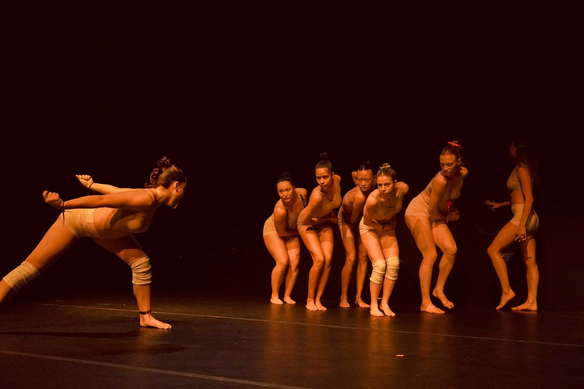 Group of women in beige dance costumes performing on stage with a dark background.