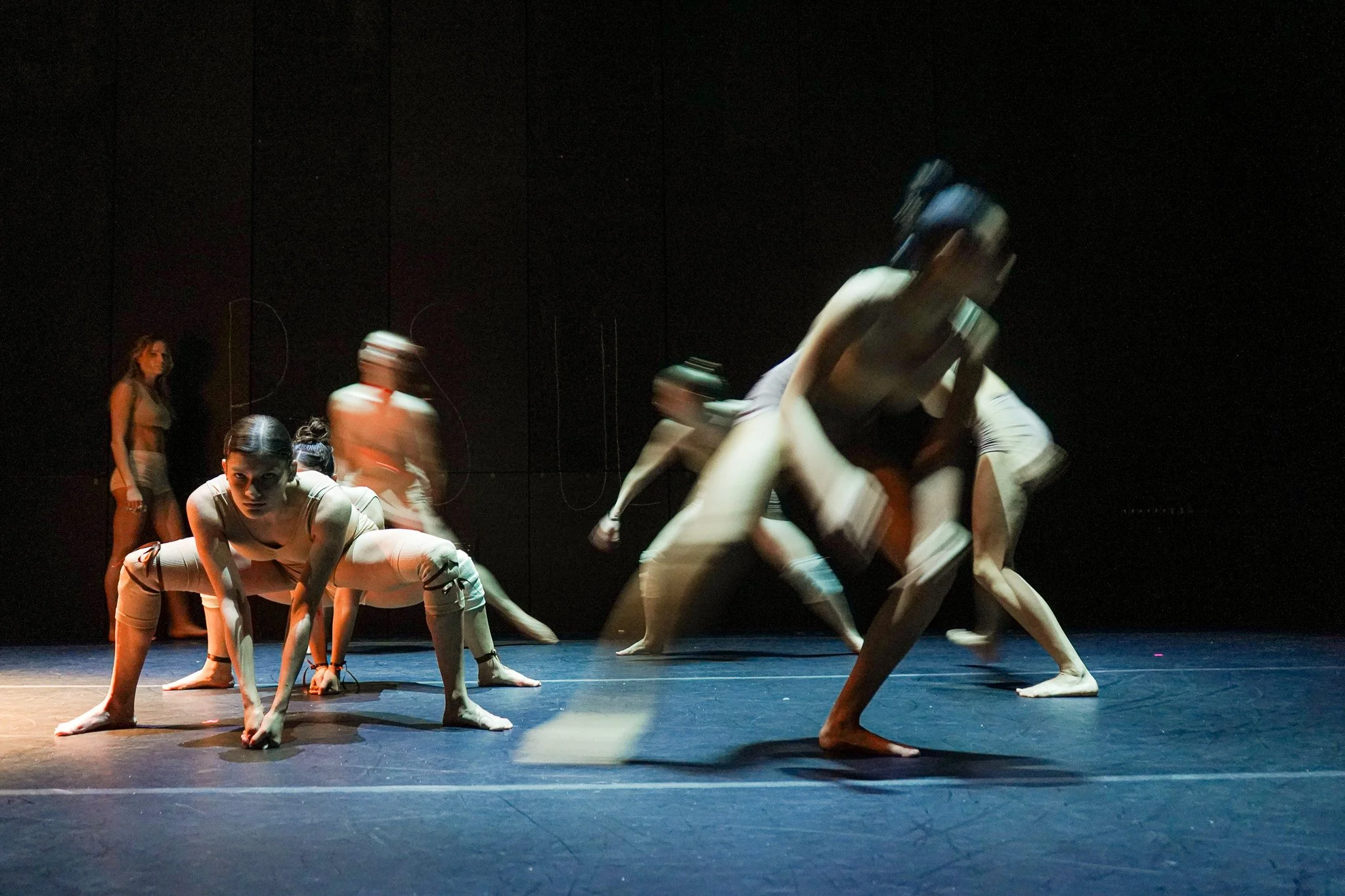 Dancers performing on stage with dramatic lighting, some crouching in the foreground and others blurred in motion.