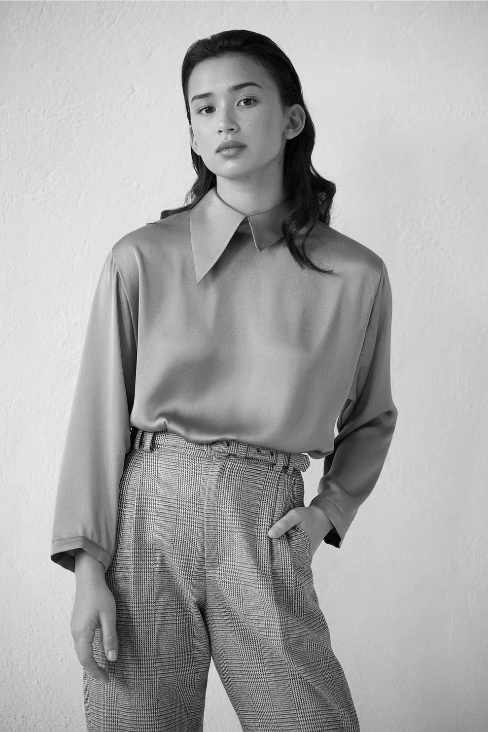 A woman in a silky shirt and patterned trousers standing against a plain wall in a black-and-white photo.