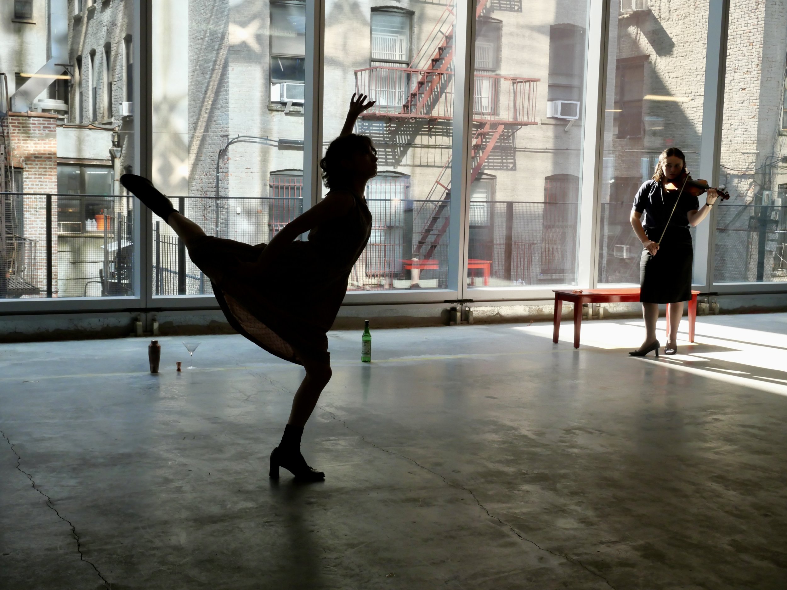 Silhouette of a woman dancer performing a pose in front of large glass windows with a musician playing violin outside.