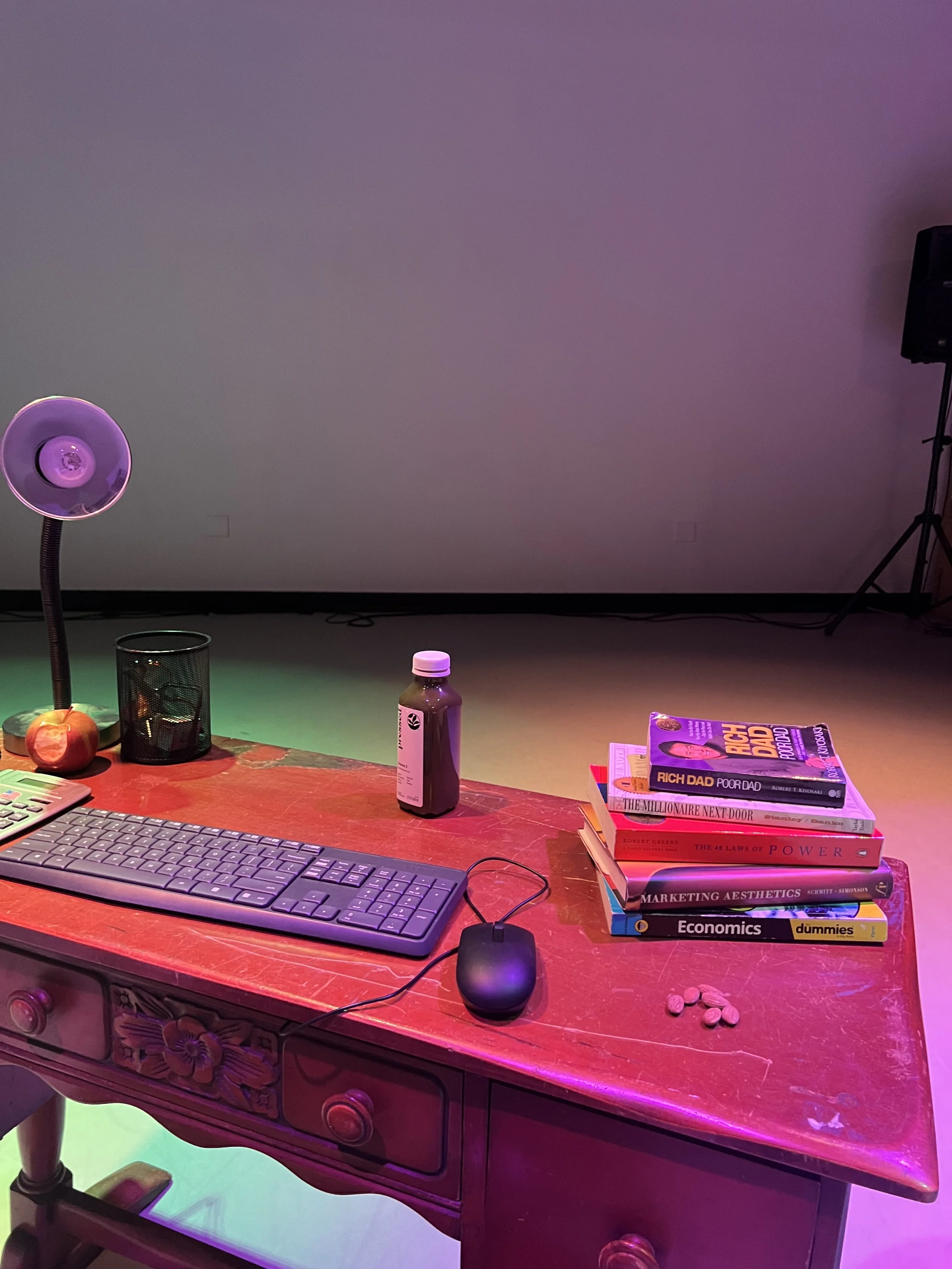 A wooden desk with a computer keyboard, mouse, and several books stacked on the right. A purple desk lamp, a bottle, and a black cup are also on the desk. Peanuts are scattered on the right side of the desk, with colorful lighting creating a vibrant 