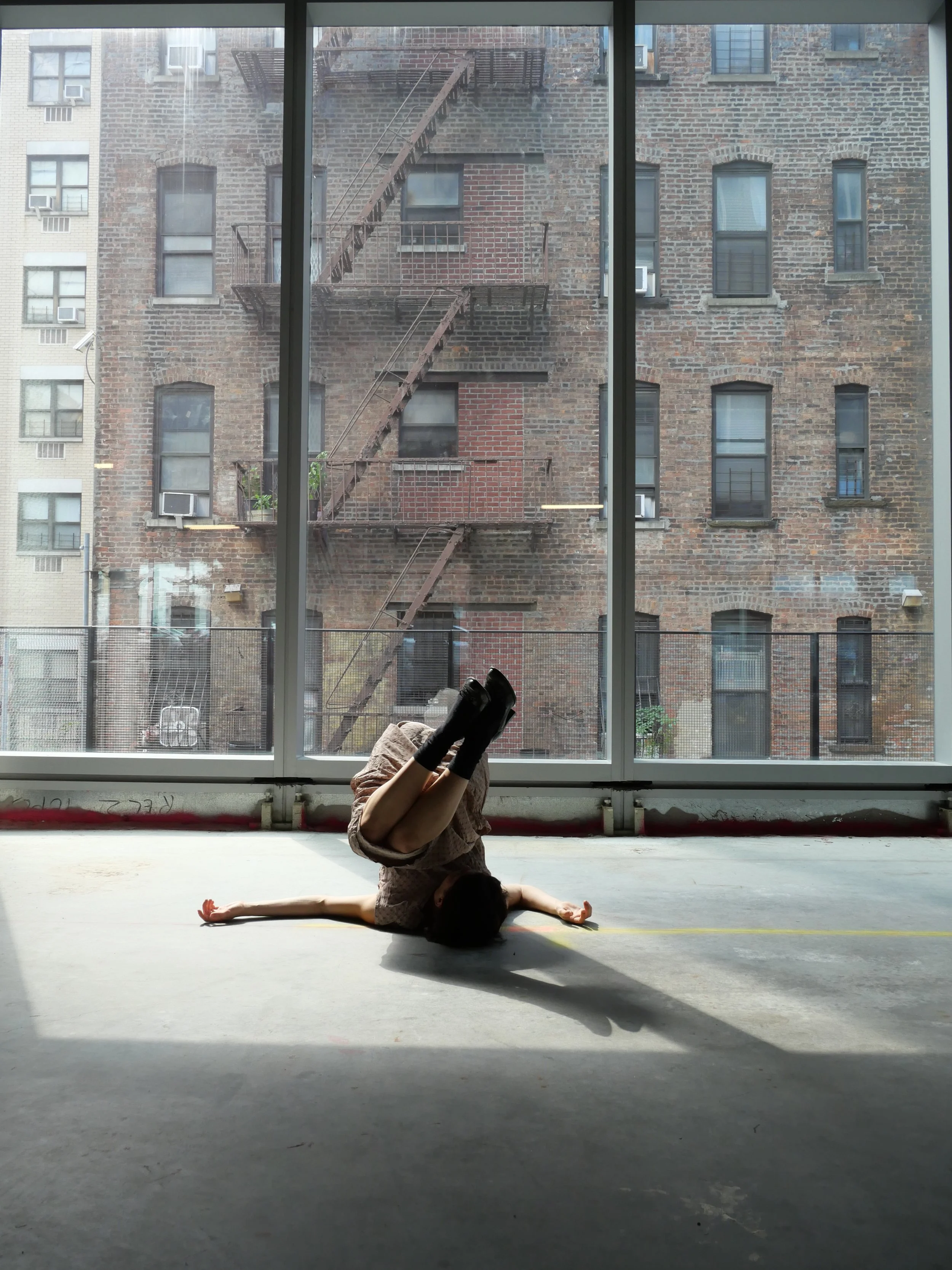 Person doing a headstand yoga pose on the floor indoors with large windows showing brick apartment buildings outside.
