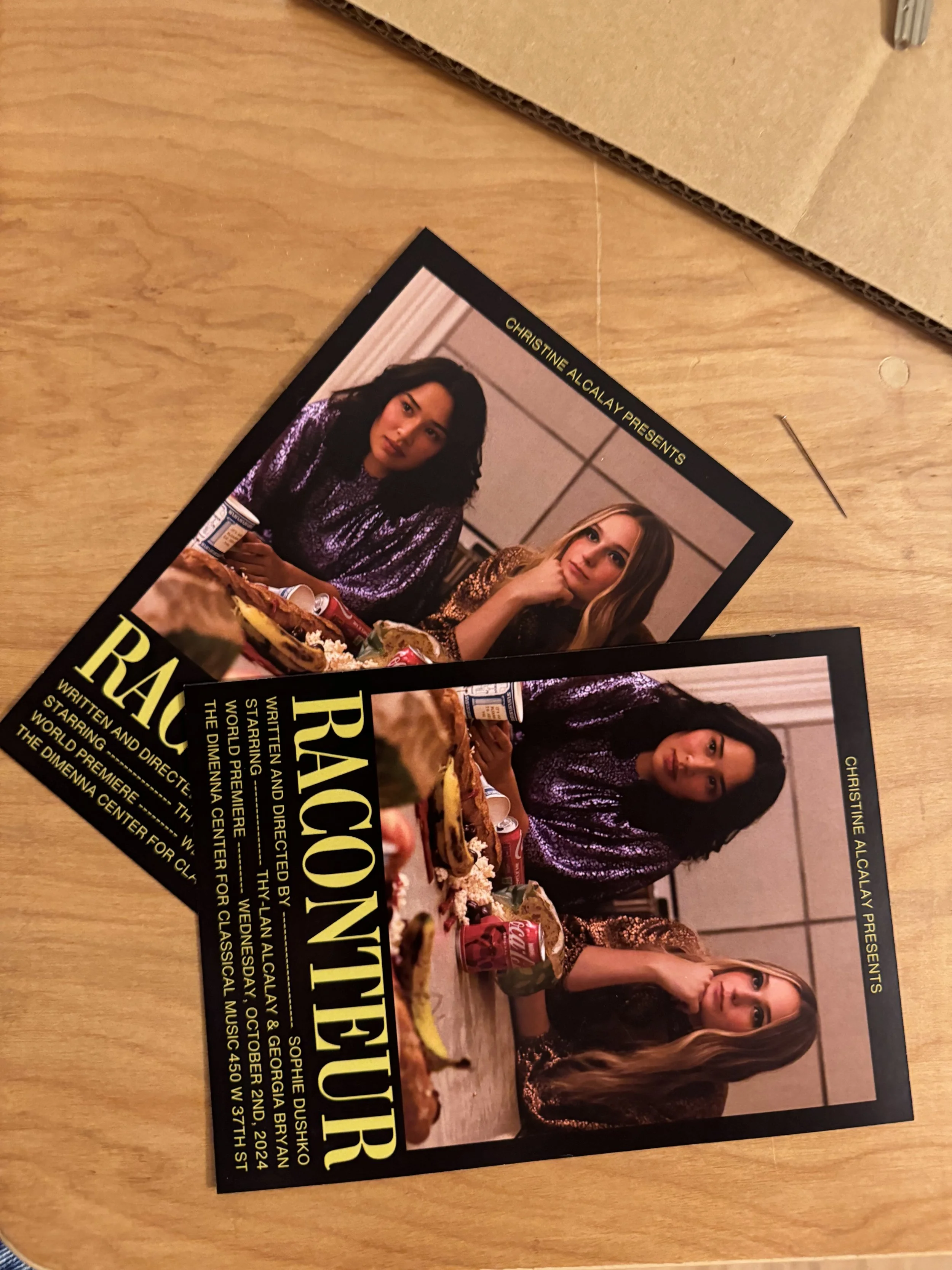 Two identical event posters on a wooden table, featuring three women at a table with food, with text about a performance by Raconteur.