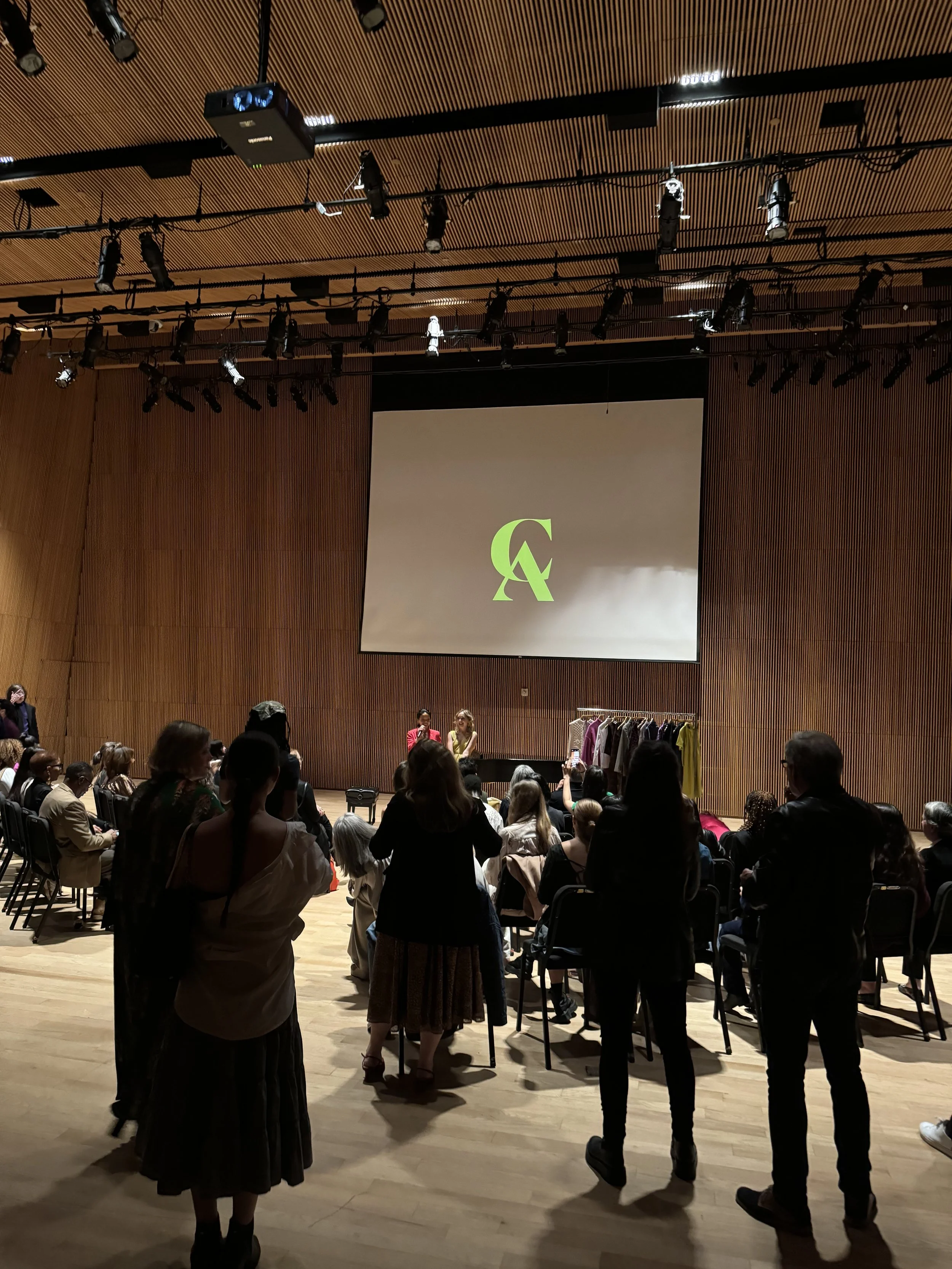 People gathered in a wood-paneled auditorium facing a stage with a large screen displaying a green letters logo.