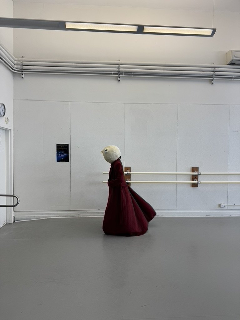 A person in a red dress with a large, round, white head walking in a plain, white room with ballet barres.