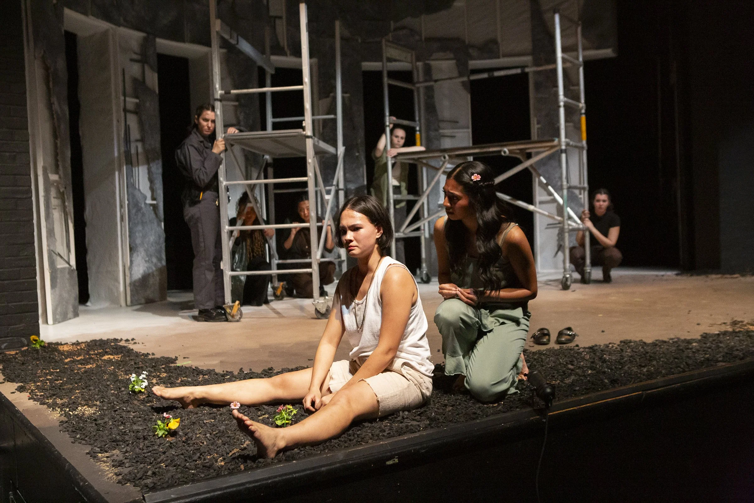 Two women are seated on a stage, with one woman sitting on the ground with her legs extended and barefoot, and the other woman kneeling beside her, slightly leaning forward and engaging in conversation. In the background, several crew members are wor