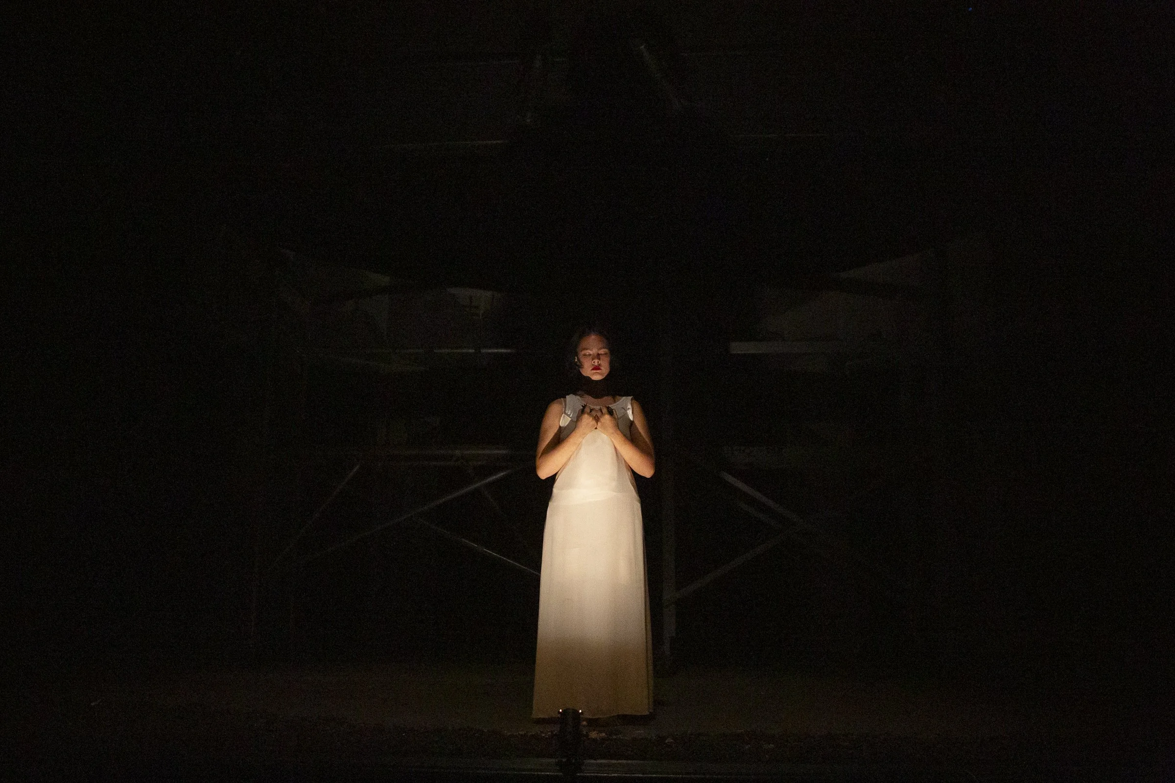 Woman in a white dress standing in darkness, holding her hands near her chest, illuminated by a spotlight.