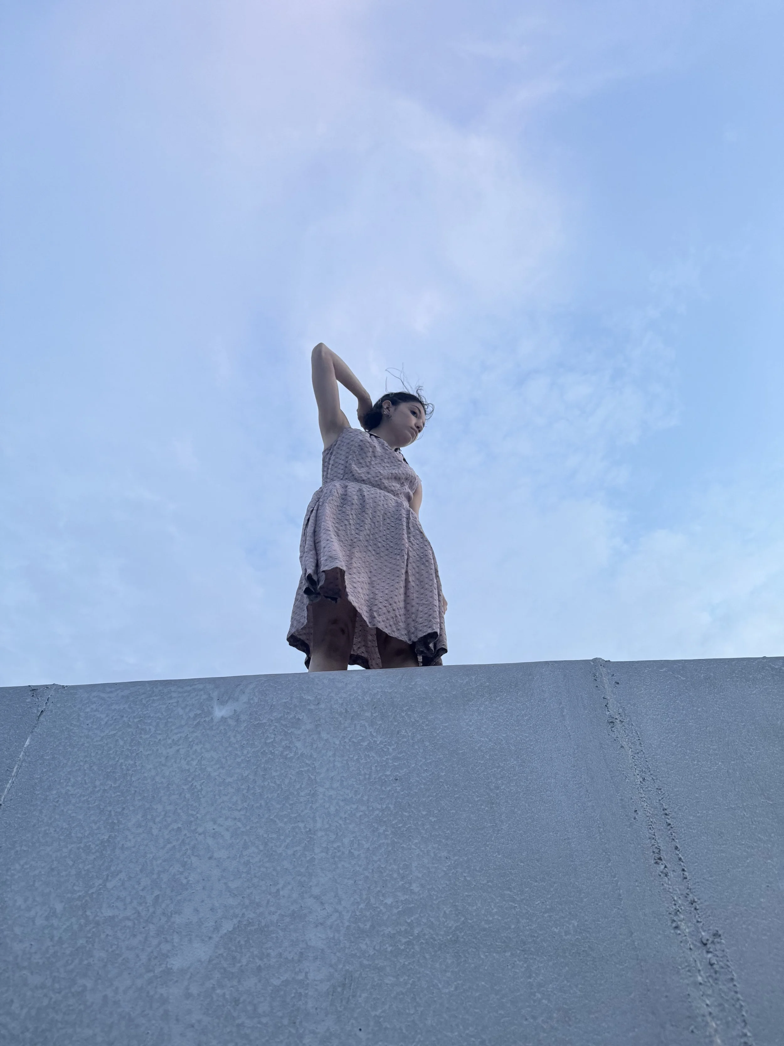 A woman standing on top of a concrete structure against a partly cloudy sky, wearing a light-colored dress and looking to the side.