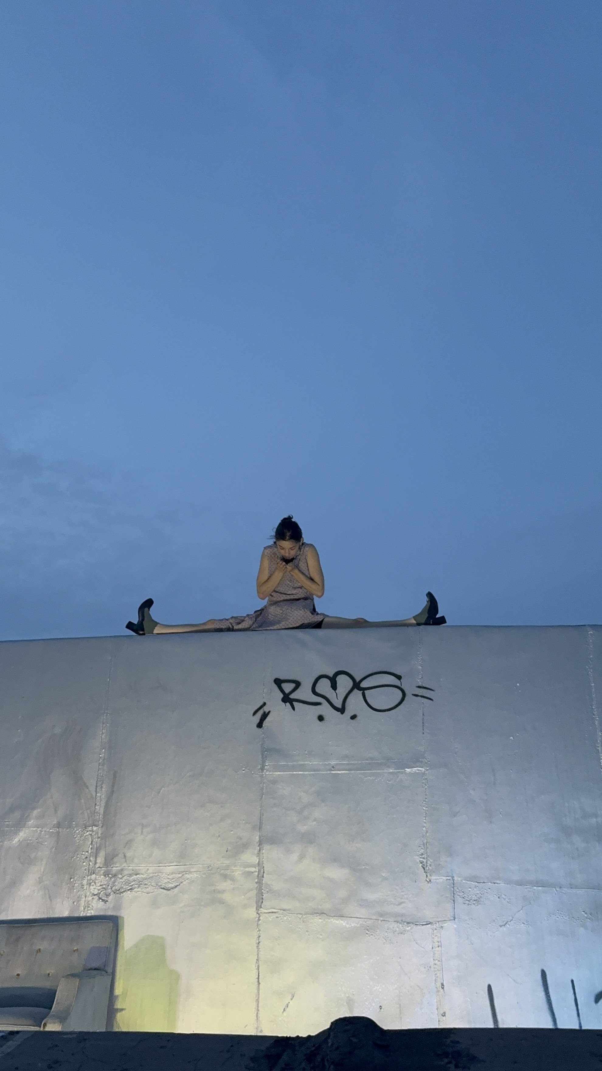 A woman sits in a split position on top of a structure, with her legs extended to the sides, while looking down and holding her hands close to her chest. The sky is visible in the background, and the structure has graffiti that says 'R♥S'.