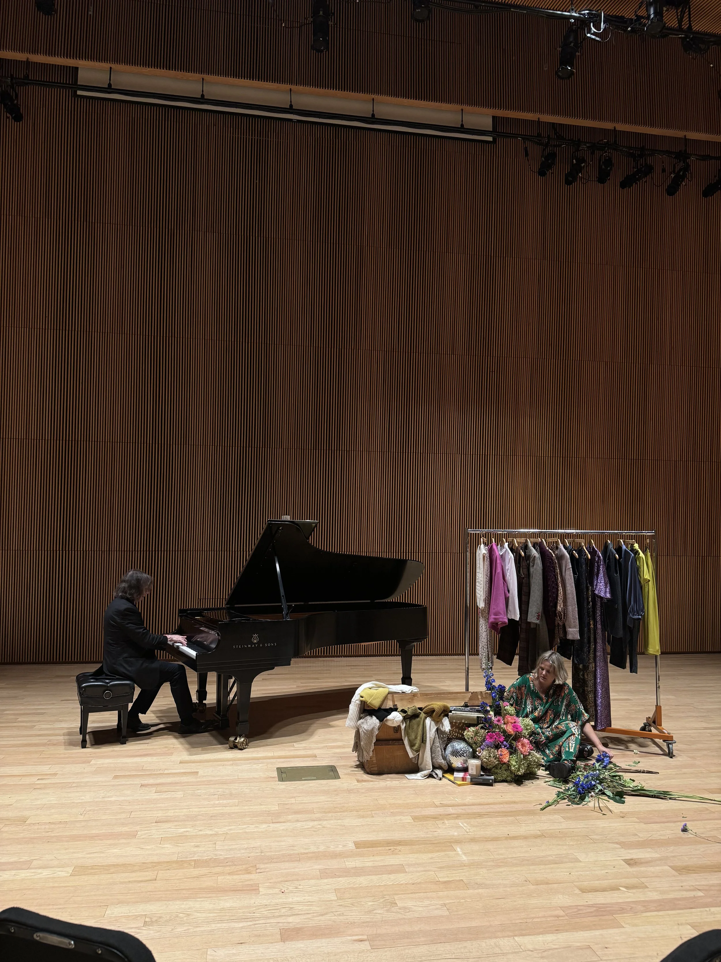 Musicians playing a grand piano on stage with a clothing rack, flowers, and miscellaneous items as background.
