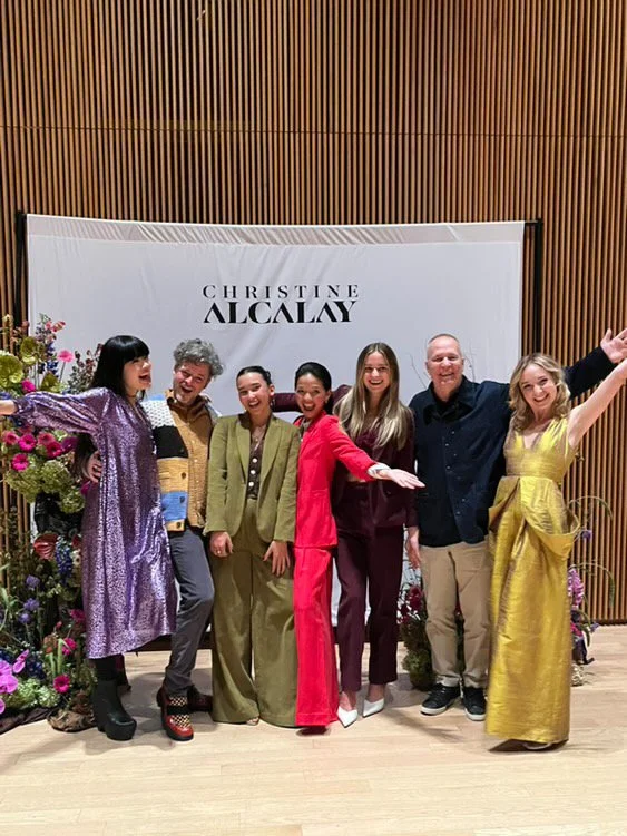 Group of seven people standing in front of a backdrop that says "Christine Alclay", smiling and posing for a photo at an event, with flowers on the left.