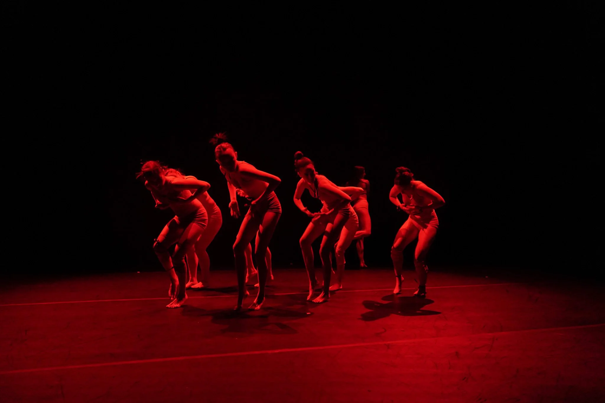 A group of dancers performing on stage in red lighting, wearing similar outfits, and striking dynamic poses.