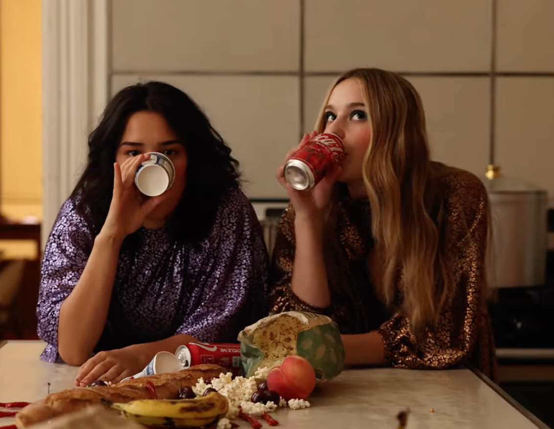 Two women sitting at a table drinking from cans, with food and snacks including a banana, apple, popcorn, and a cake on the table.