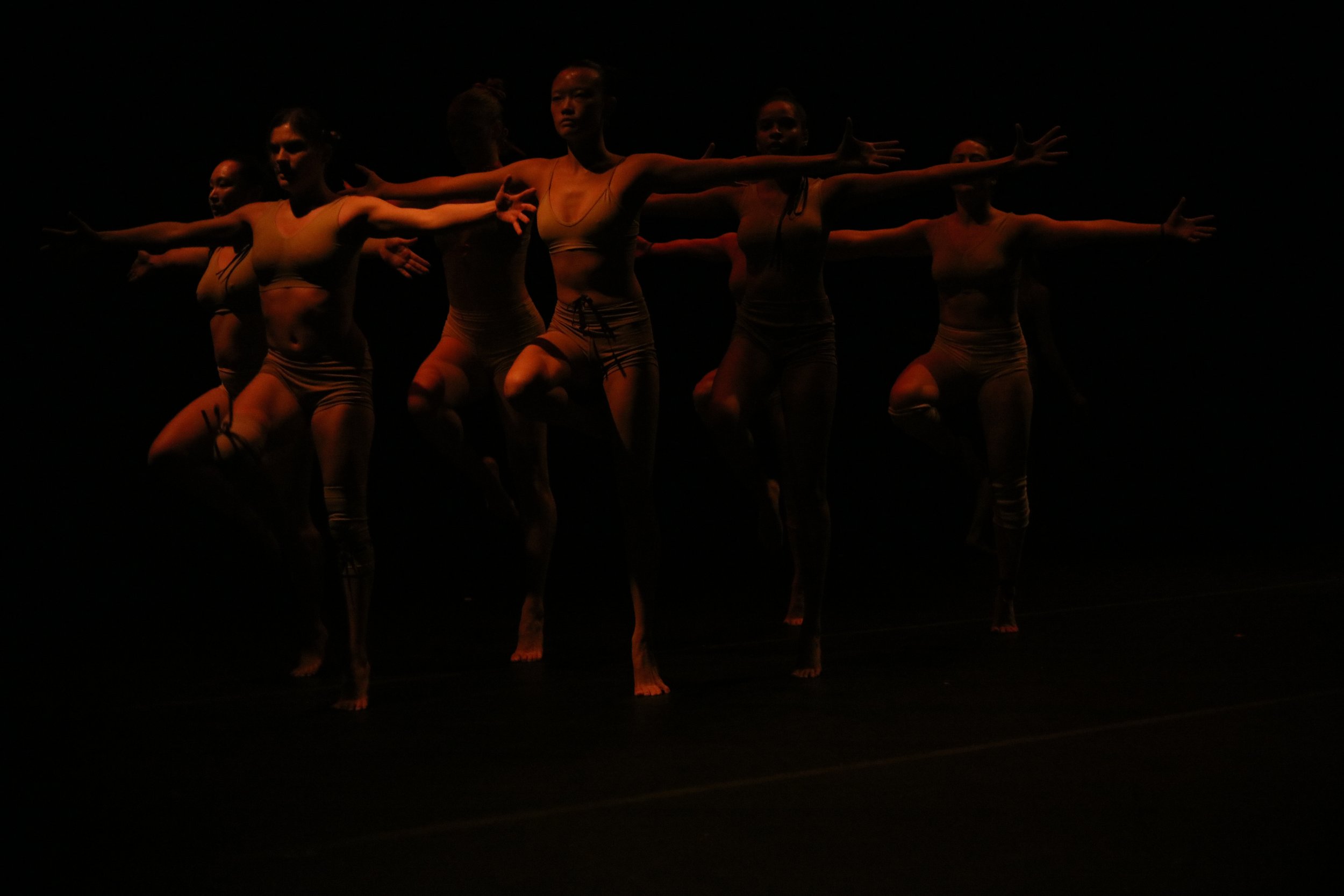 A group of seven dancers performing on a dark stage, with some dancers lifting one knee and extending their arms.