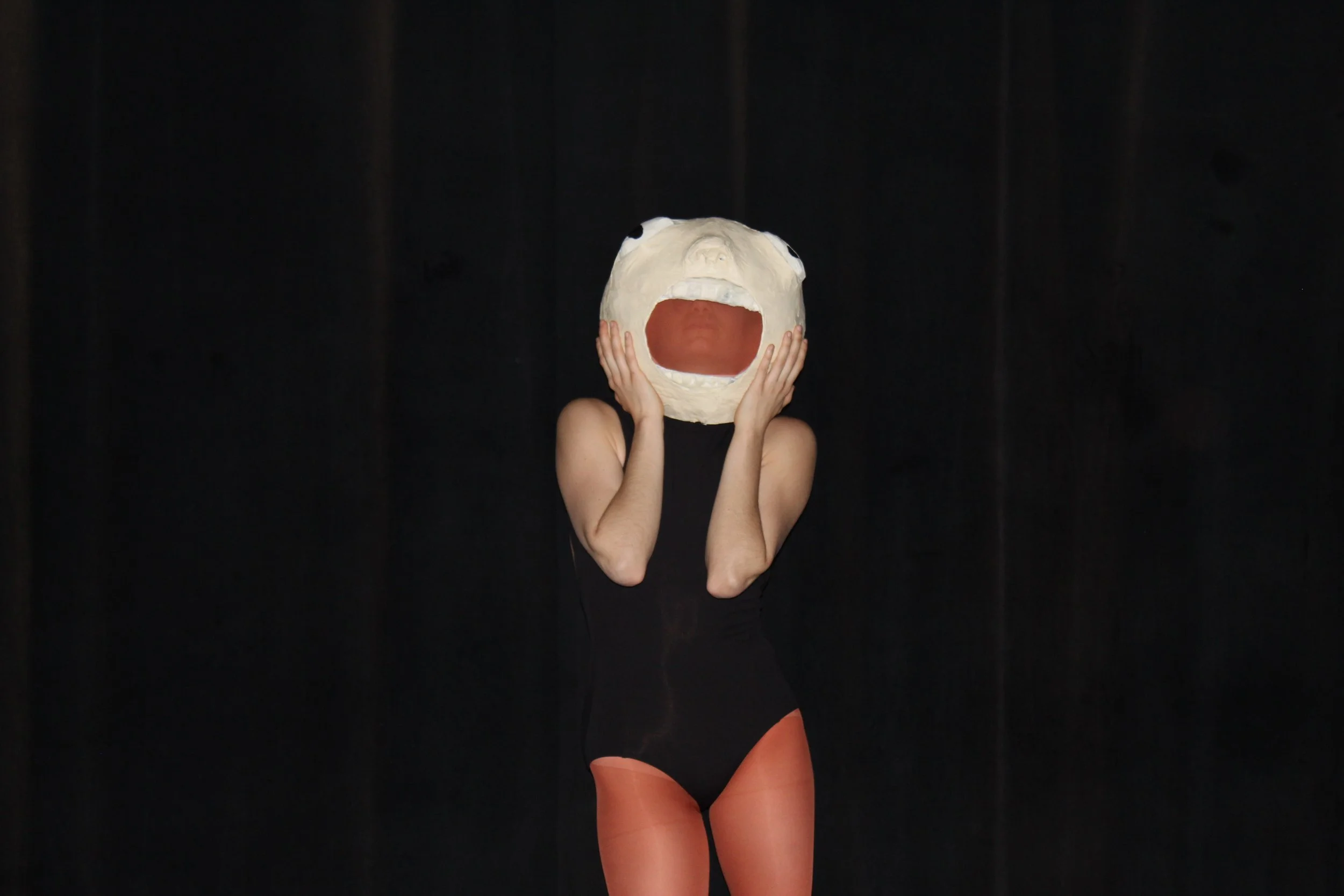 Person wearing a bear mask with mouth open, standing against a black background, dressed in black top and orange tights.