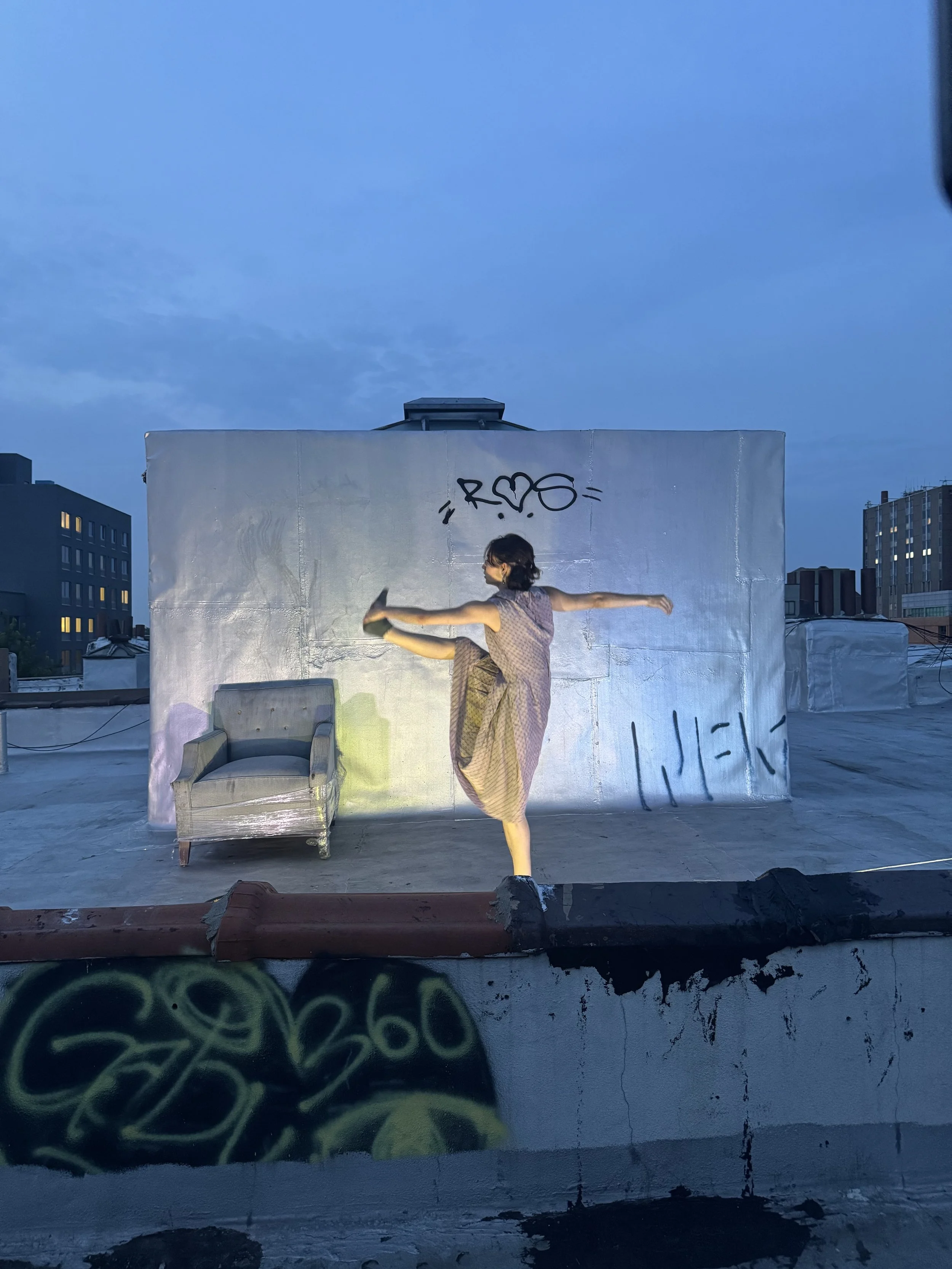 Woman practicing a high kick on rooftop with graffiti wall, chair, and city skyline in the background during dusk.