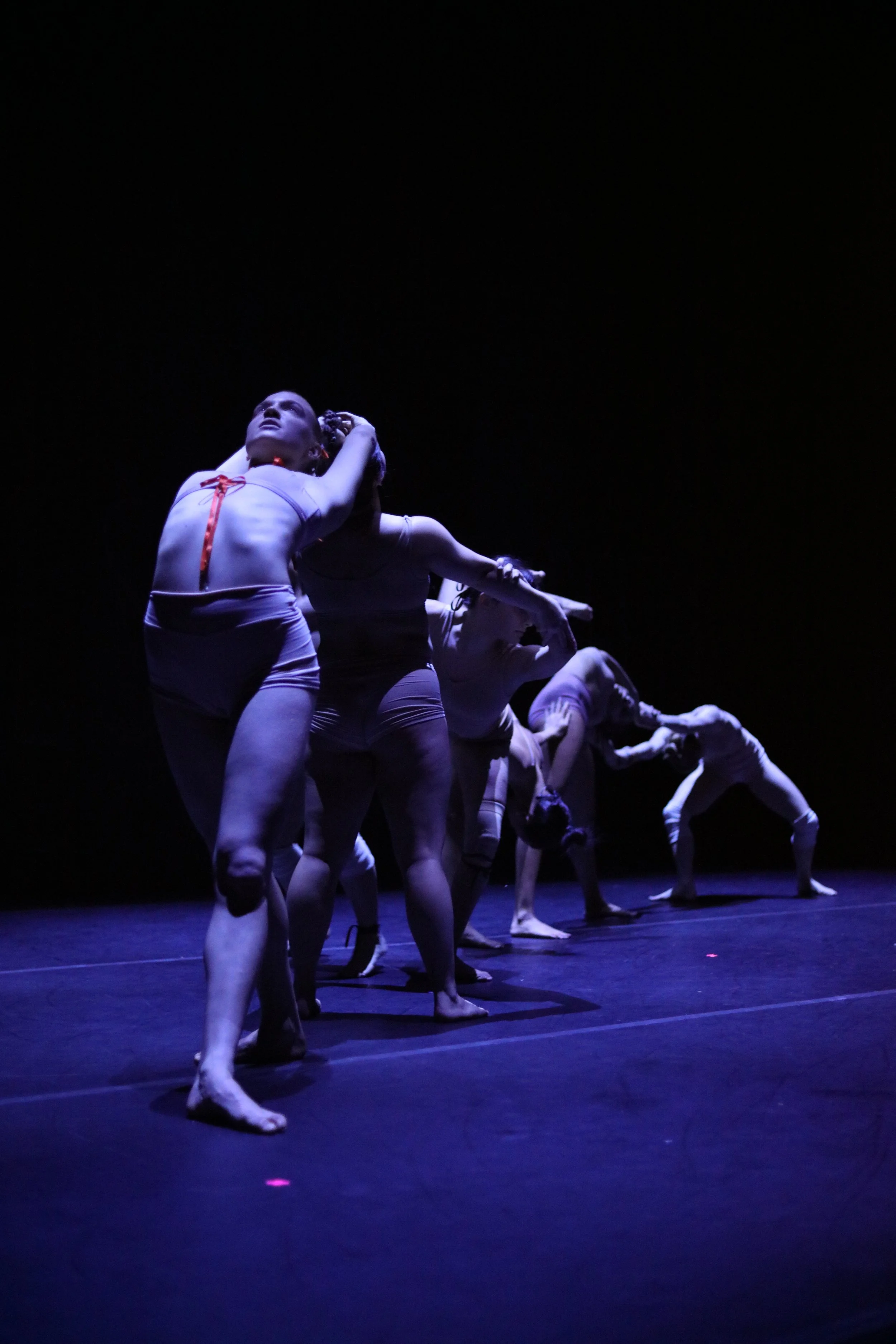 A group of dancers performing on stage under blue lighting, with one dancer in the foreground looking upward and others in various poses behind.