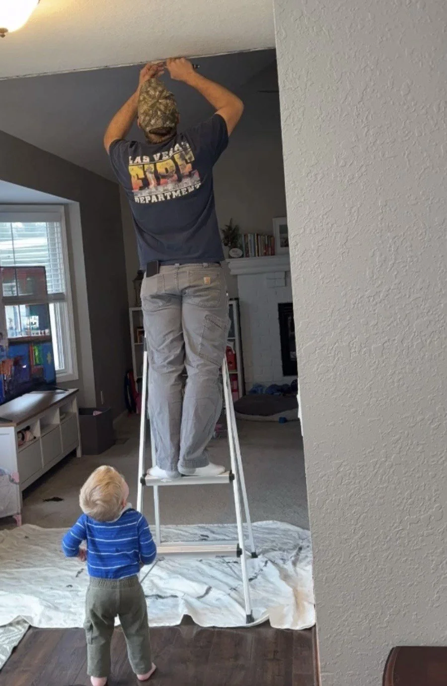 A man standing on a step ladder reaching up to the ceiling while a young child watches.
