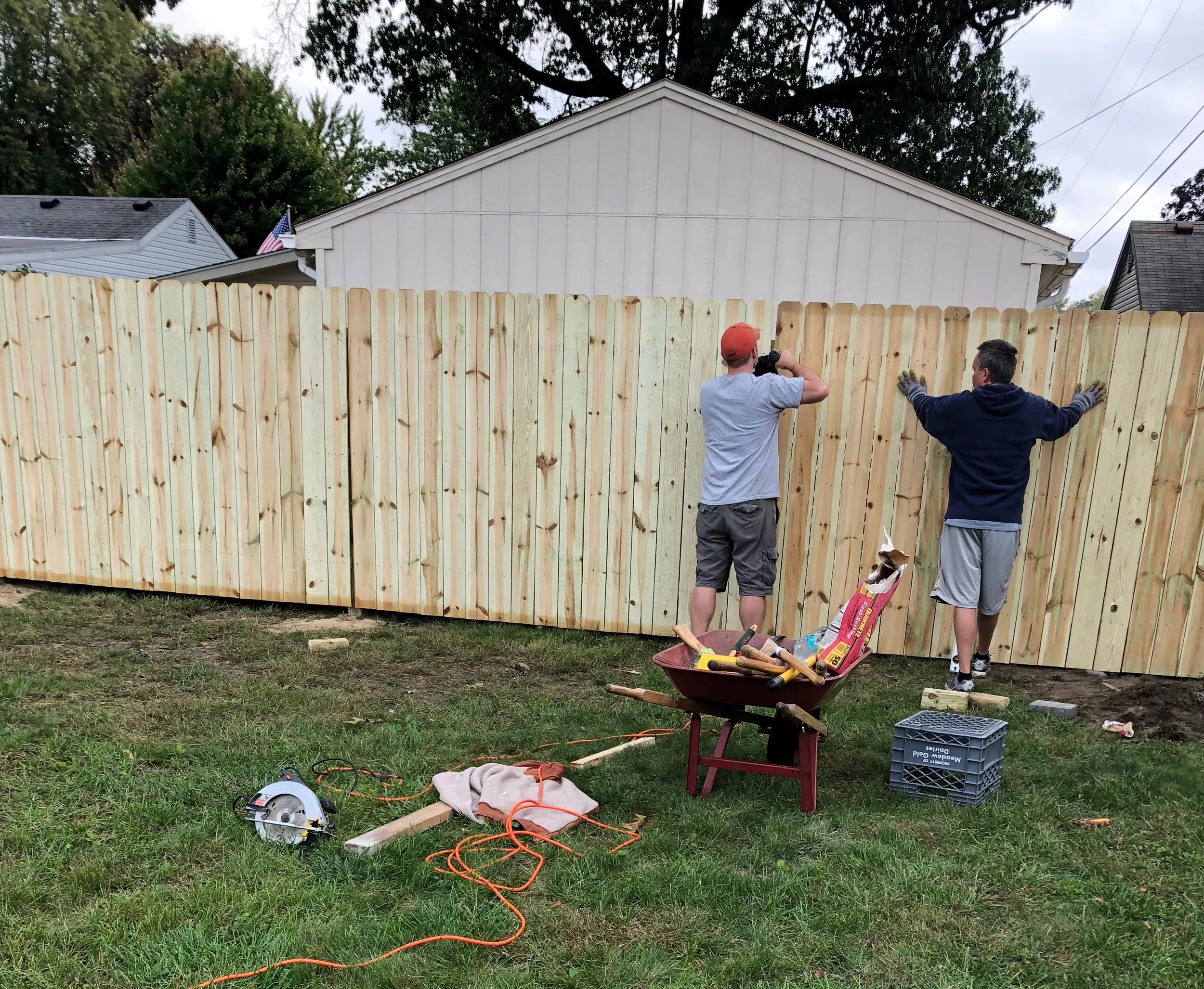 Two men installing a wooden fence in a backyard, with gardening tools and equipment scattered on the grass.