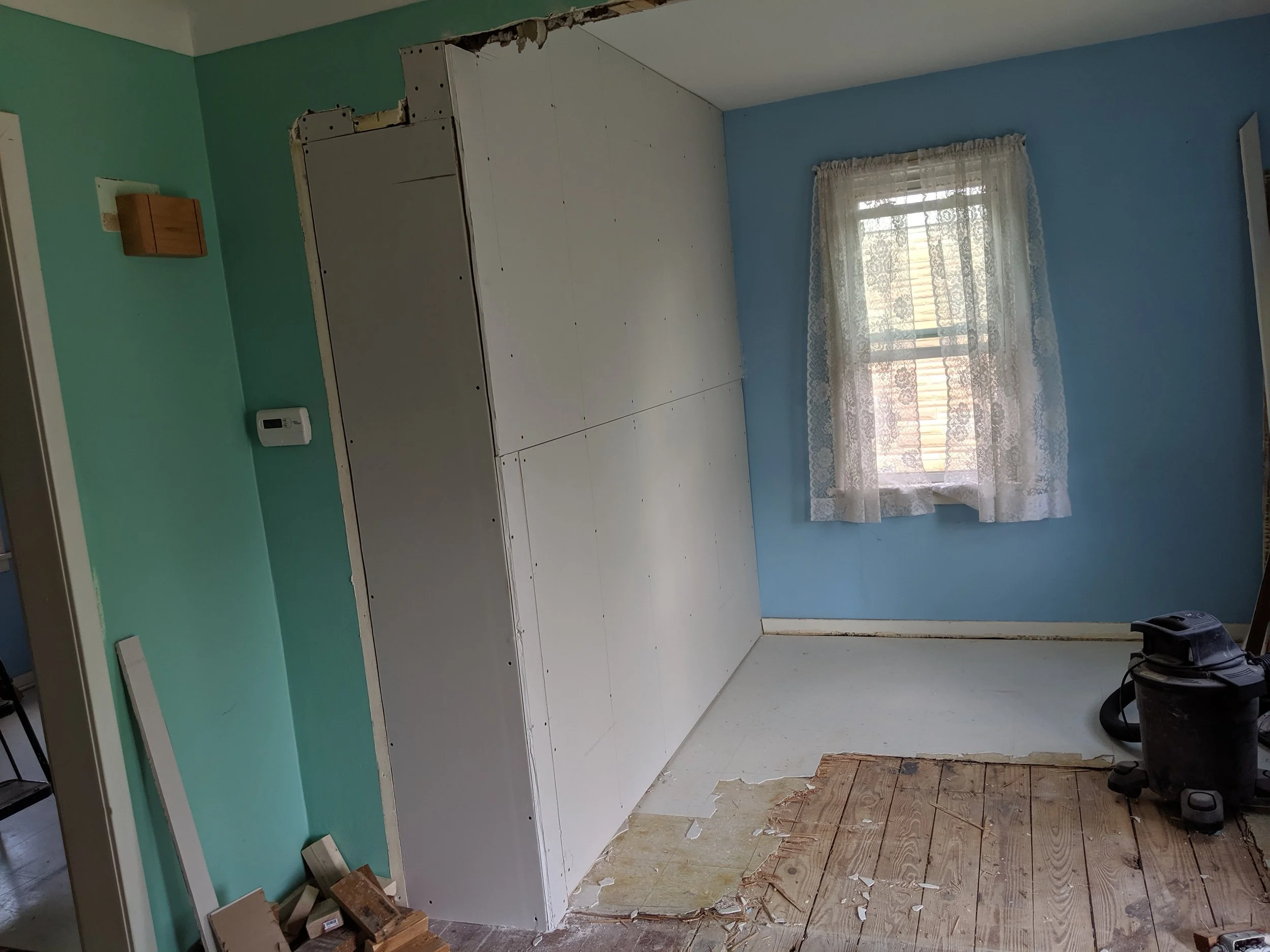 Room under renovation with partially installed wall paneling, a window with lace curtains, a vacuum cleaner, and a partially removed wood floor.