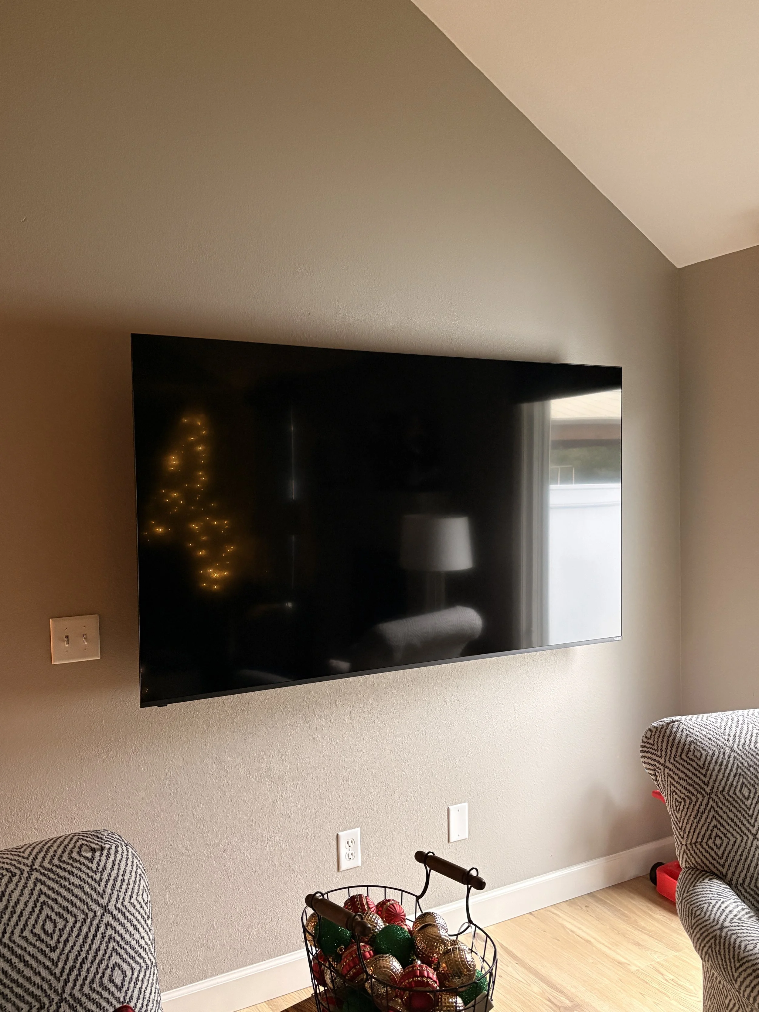 A flat-screen TV mounted on a beige wall in a living room. The TV reflects a Christmas tree with lights, a window, and a lamp. There are two chairs with patterned fabric on either side of a basket filled with Christmas ornaments on the floor.