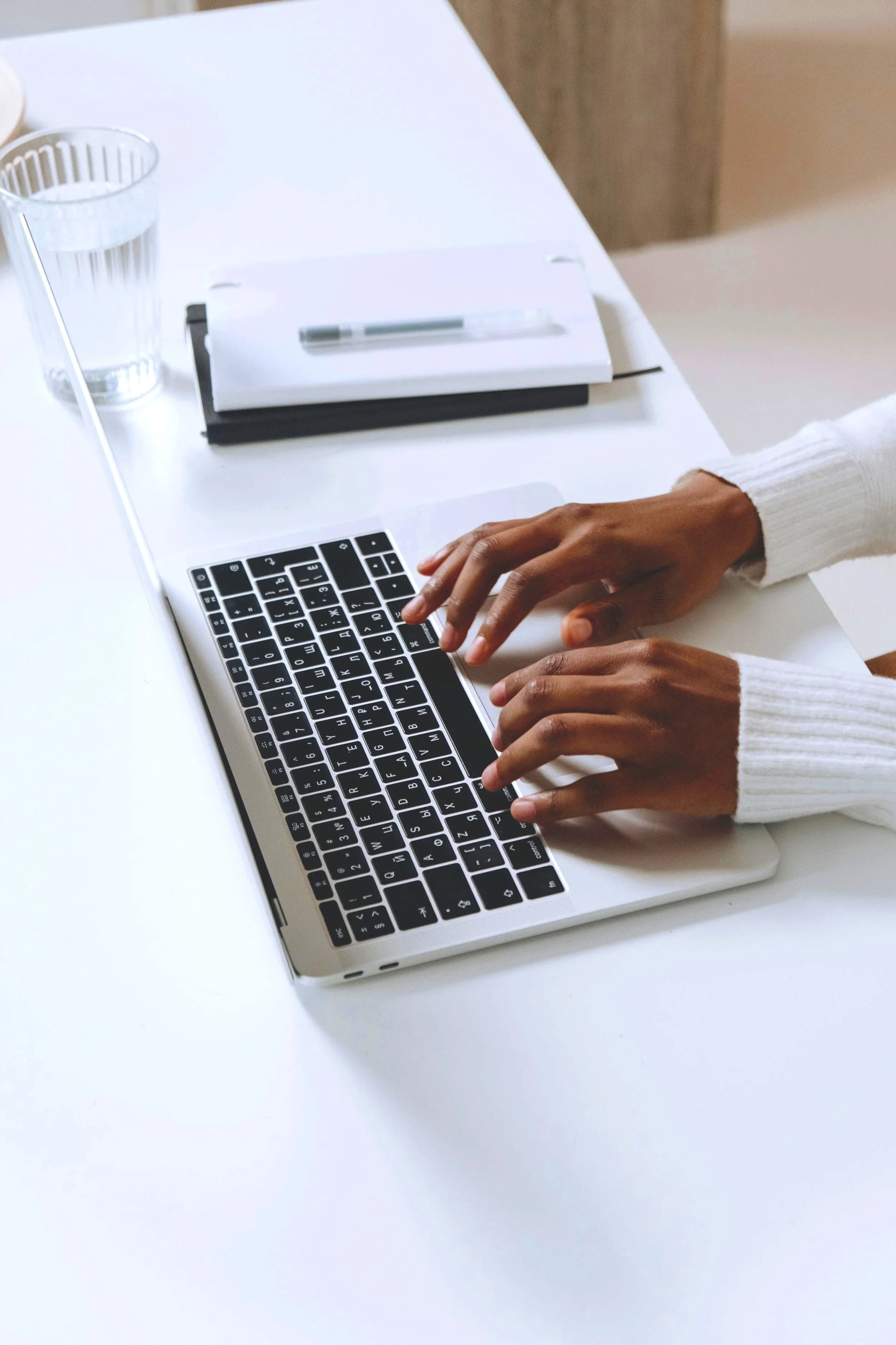 A person typing on a silver laptop at a white desk with a glass of water, a notebook, and a pen.
